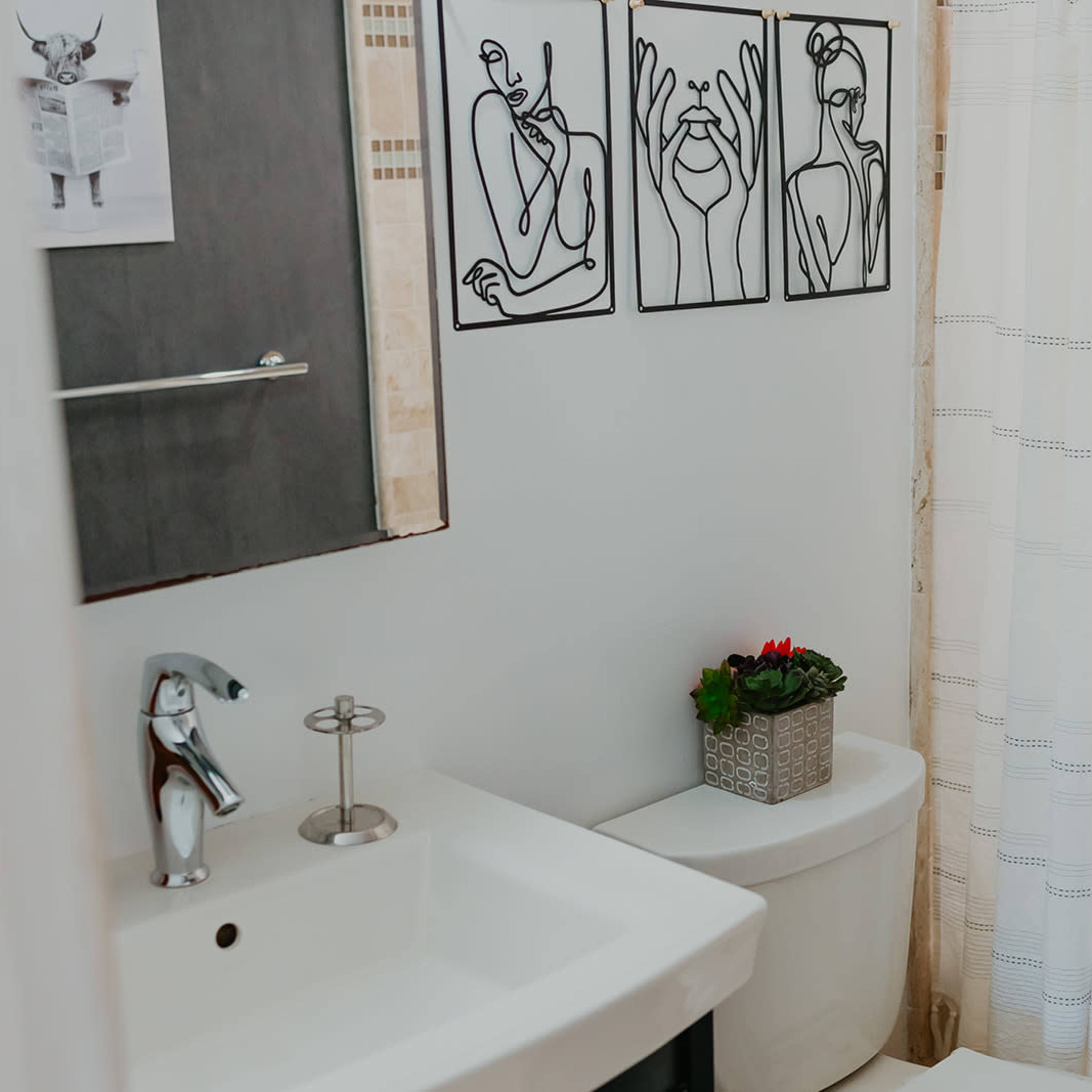 A bathroom features a modern sink, a decorative potted plant on the toilet, a shower curtain, and three abstract line art pieces on the wall.