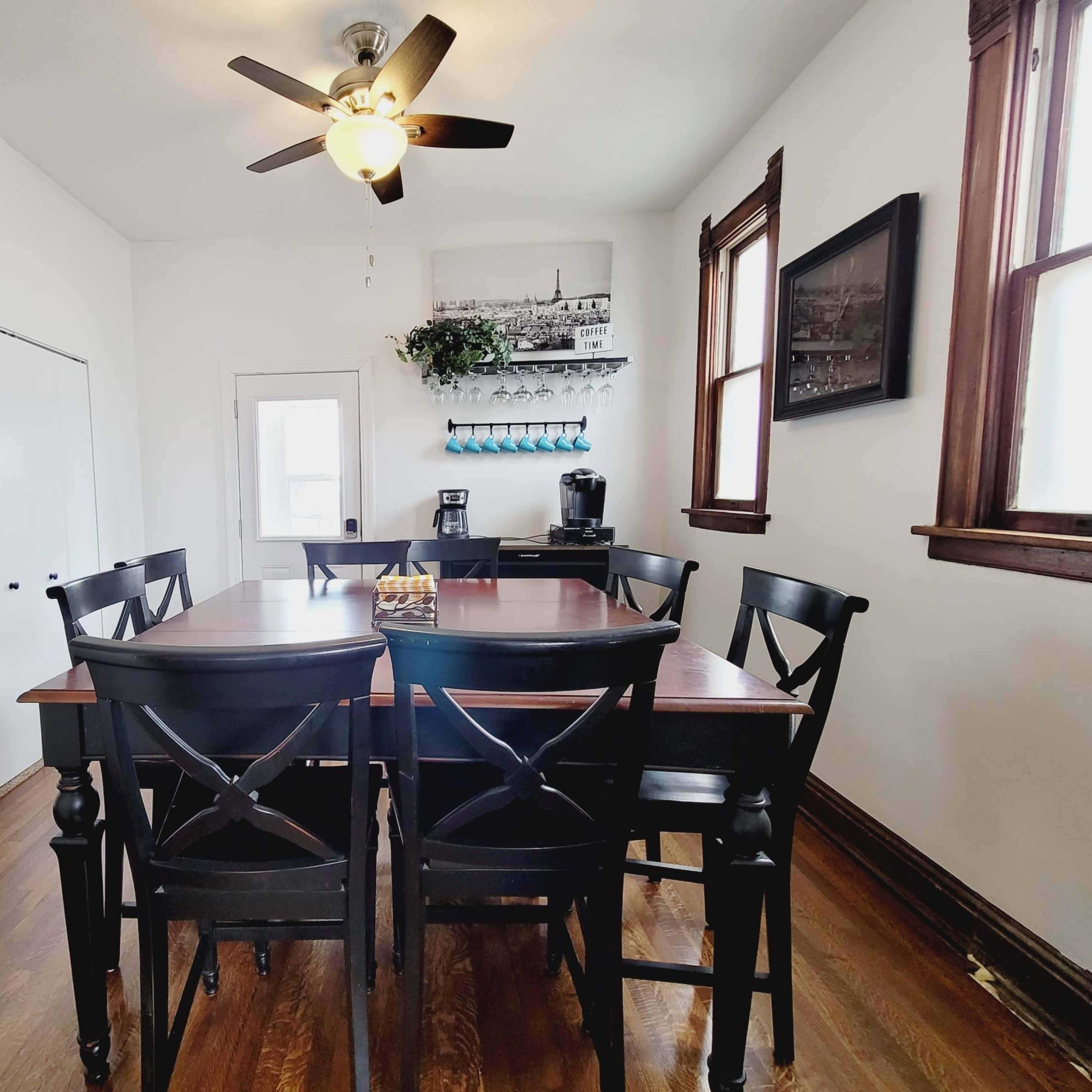 A dining area features a wooden table surrounded by black chairs and a coffee station near a door leading outside.