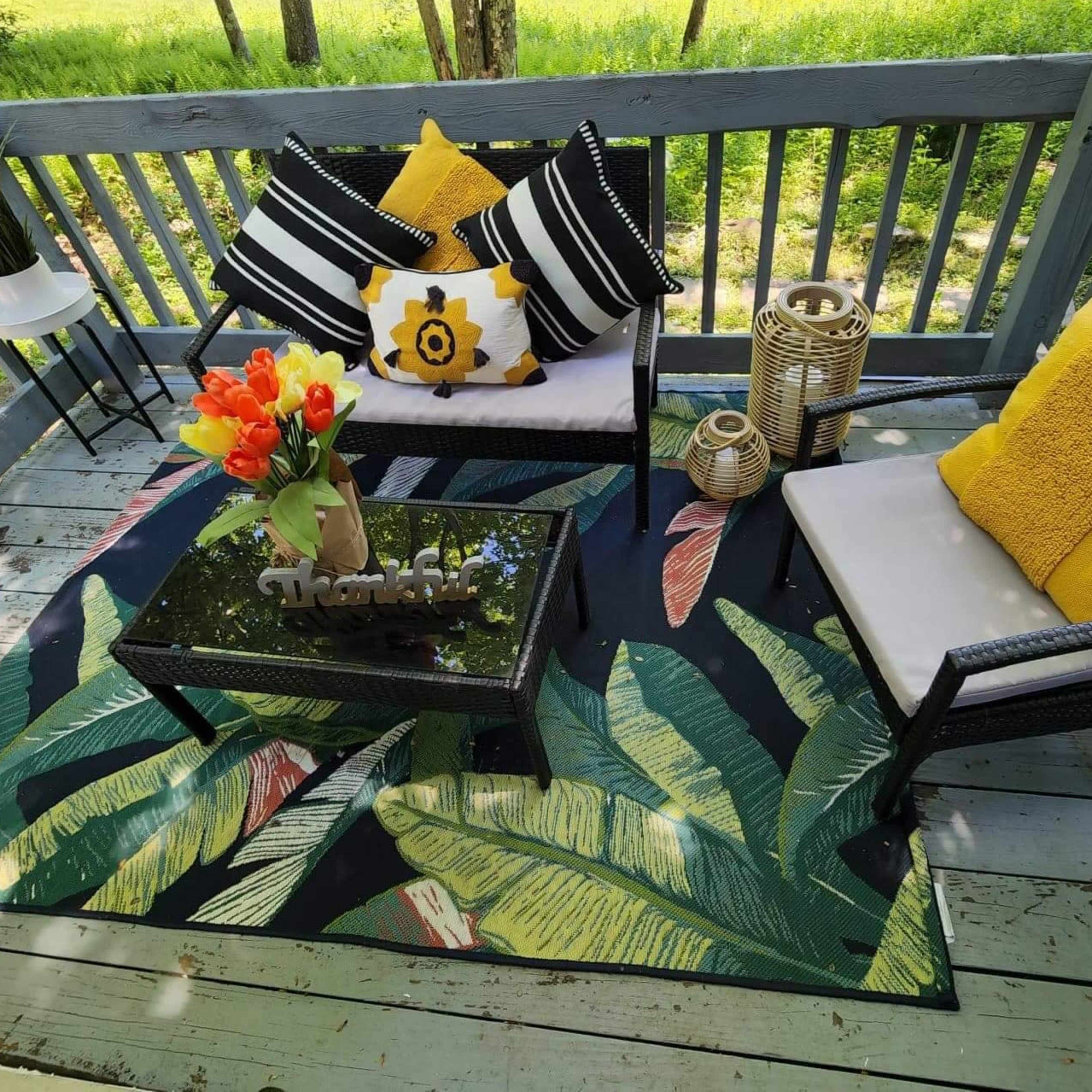 A cozy outdoor seating area features two chairs with colorful cushions, a coffee table, and a vibrant rug with tropical leaf patterns, all set on a wooden deck surrounded by greenery.