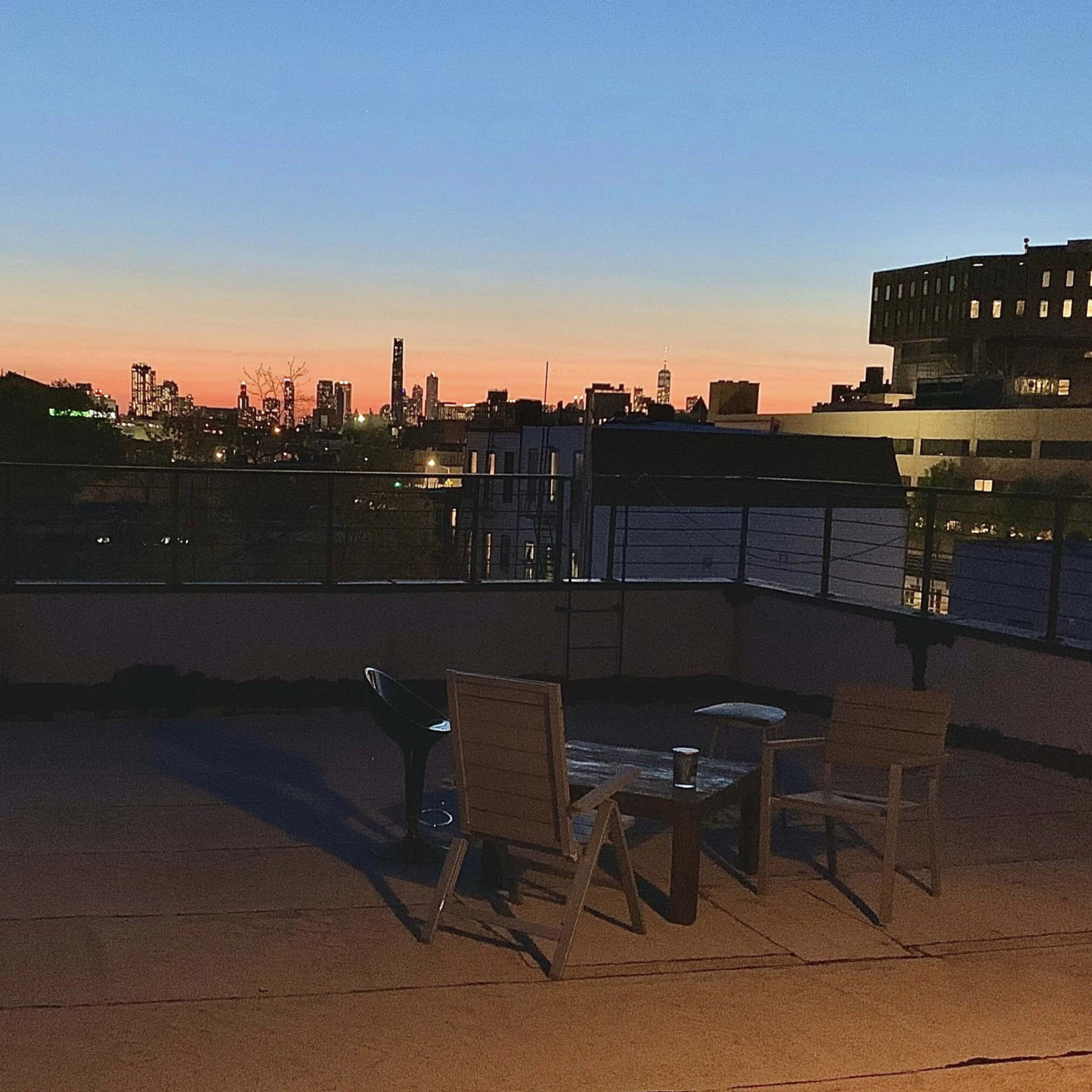 A rooftop patio with two chairs and a table overlooks a city skyline at sunset.