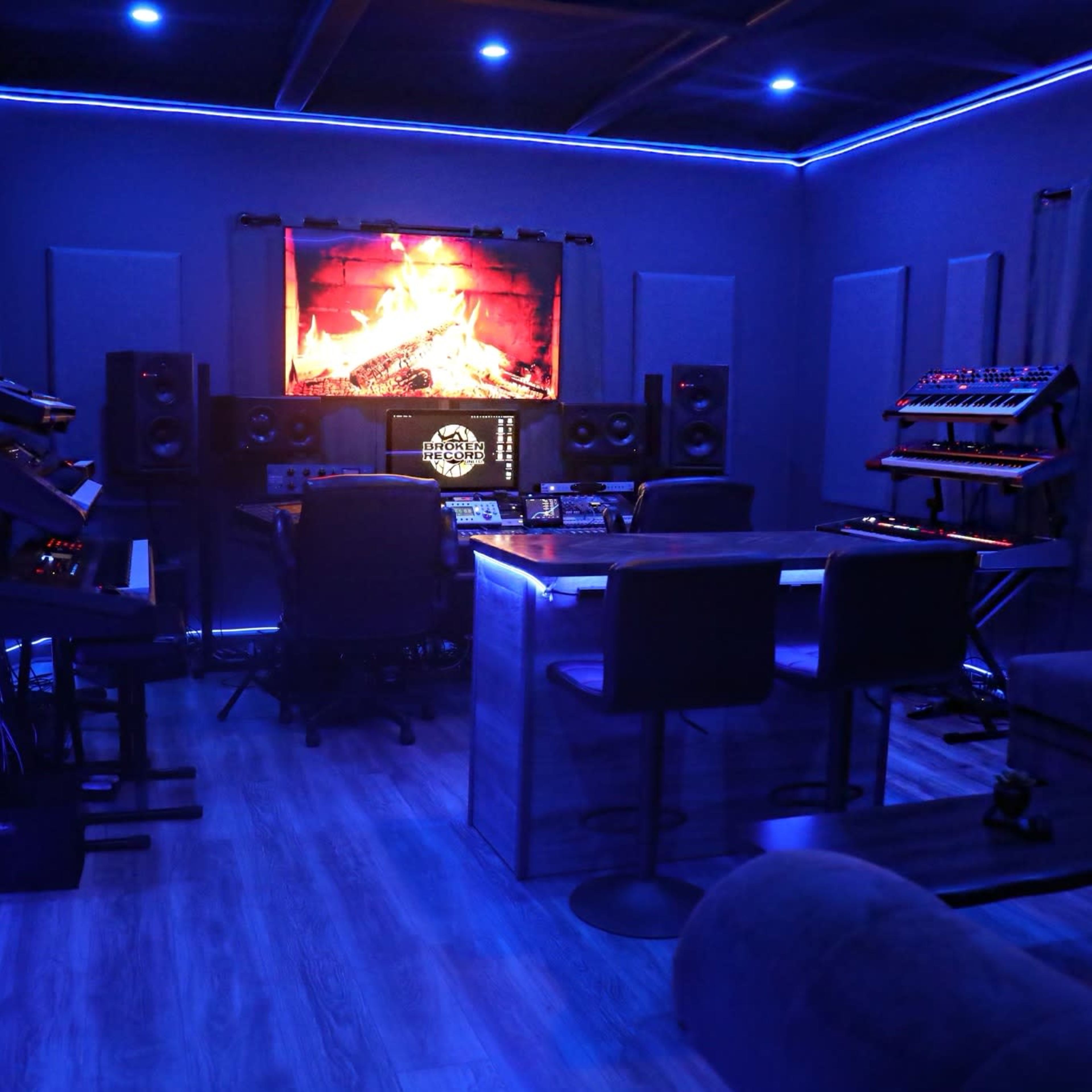 A music production studio with blue walls, a large wall-mounted screen displaying a fireplace, and various electronic instruments arranged around a central table.