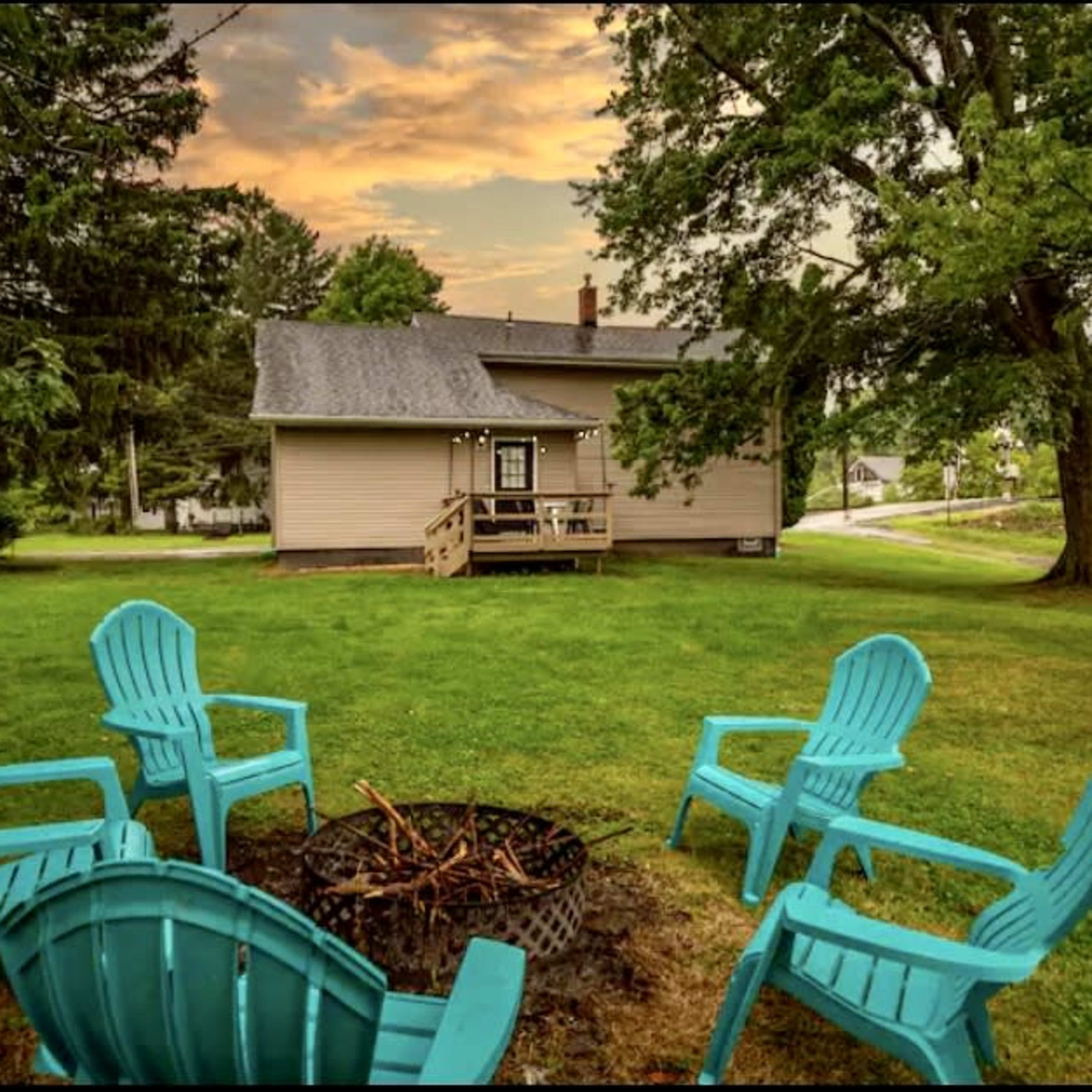 A fire pit surrounded by turquoise chairs is set in the grassy yard of a single-story house under a cloudy sky.