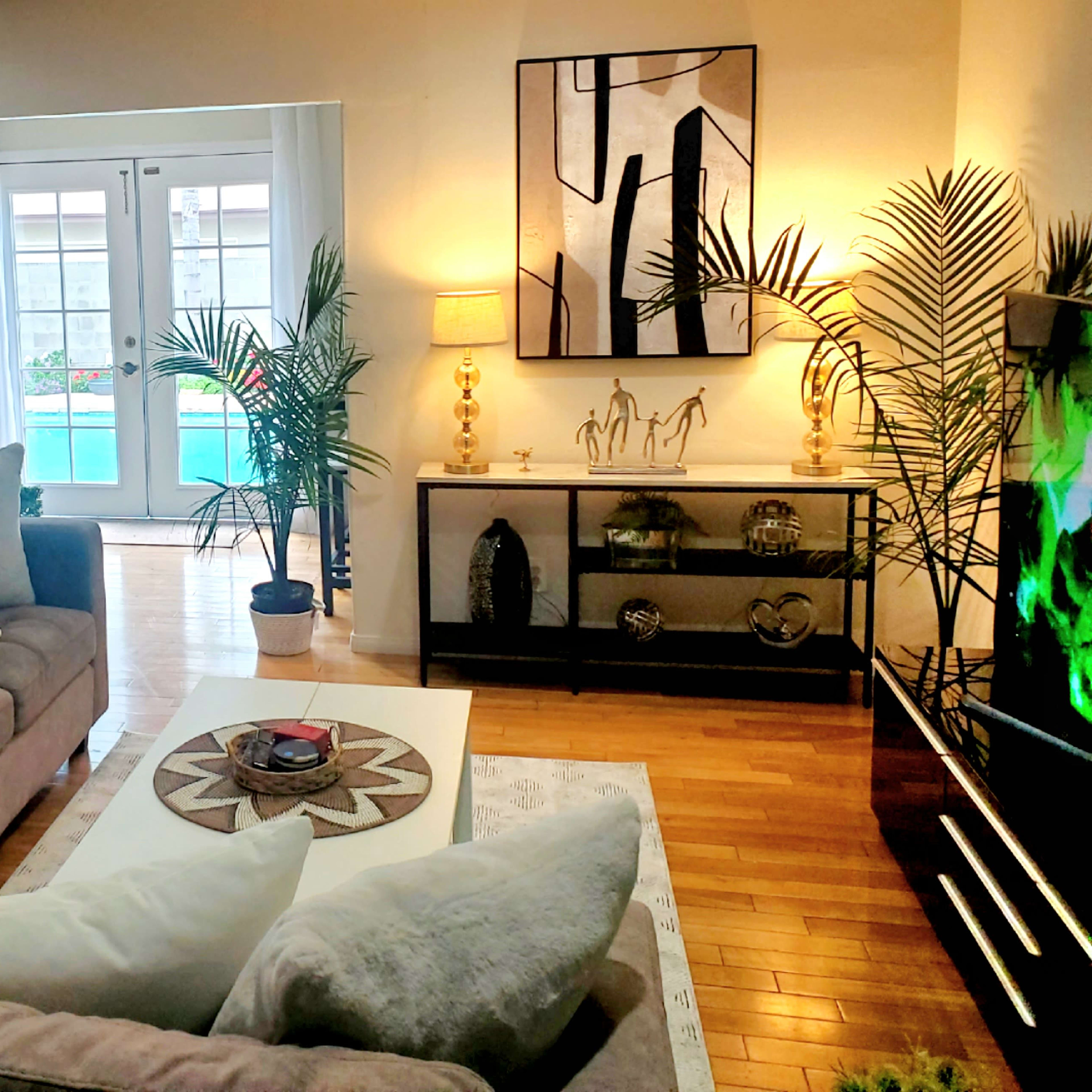 The living room features a sectional sofa, a coffee table, decorative plants, and an open view to a pool area through glass doors.
