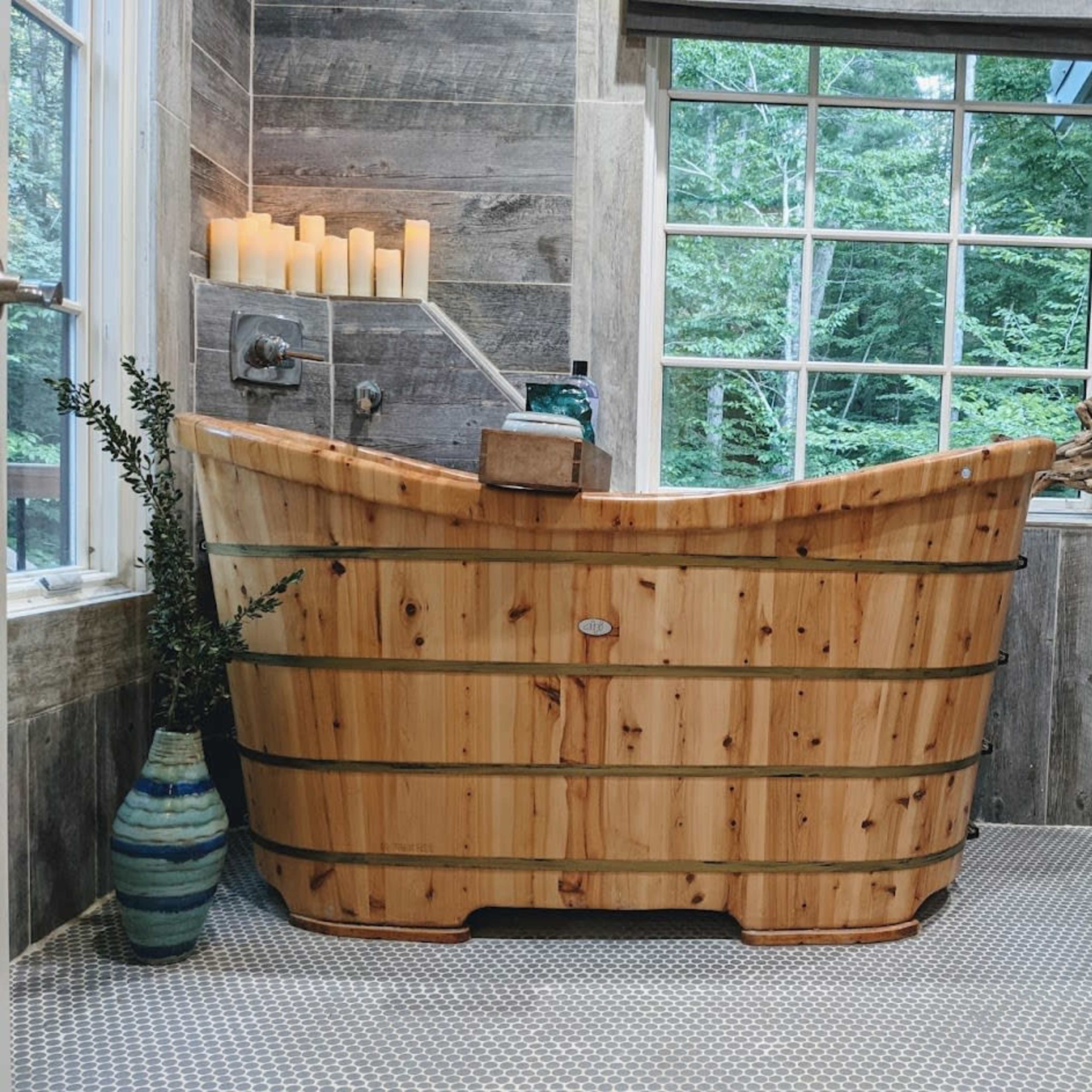A wooden bathtub is positioned in a room with stone walls and a large window overlooking greenery.