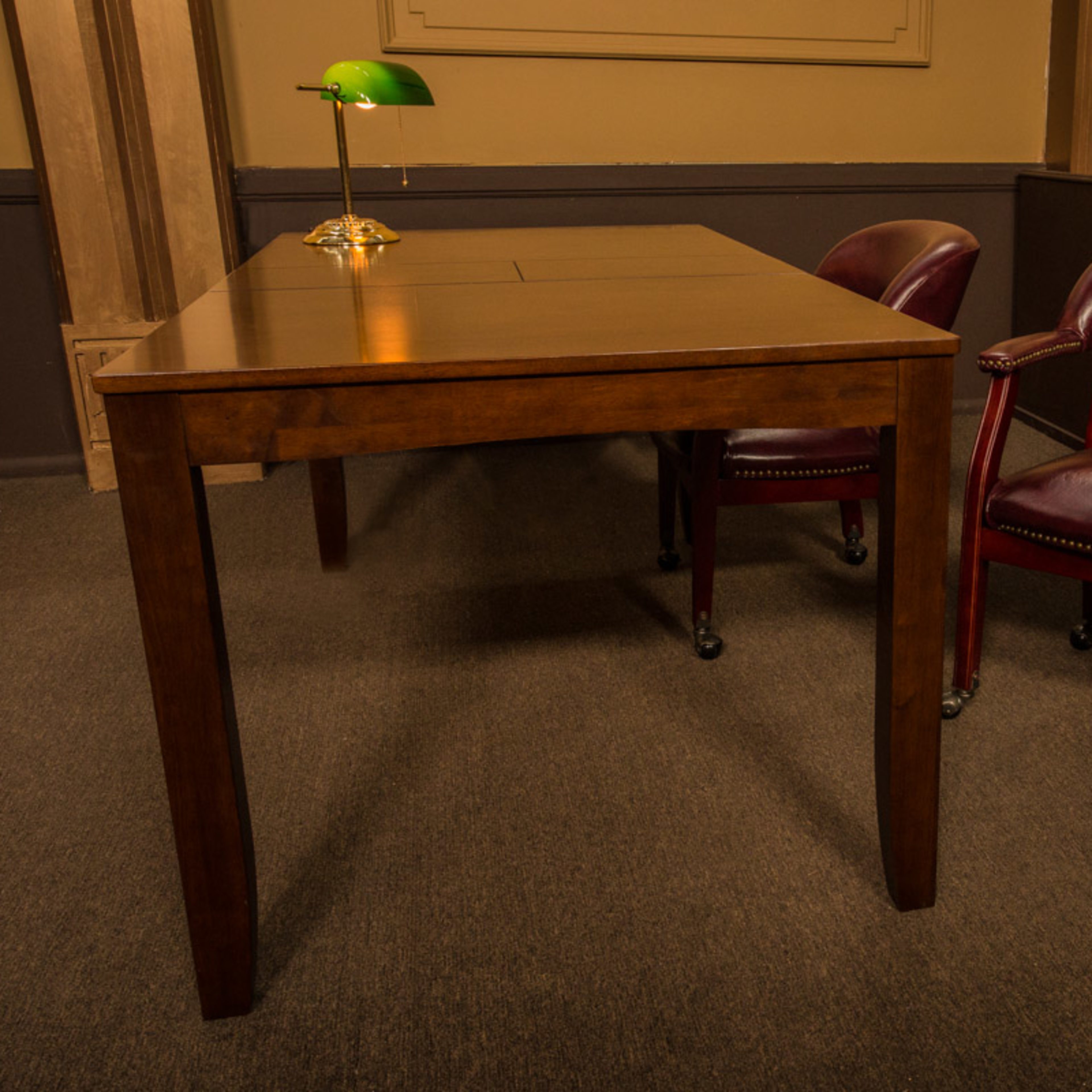 A wooden table with a green lamp sits between two red leather chairs in a room with brown walls.