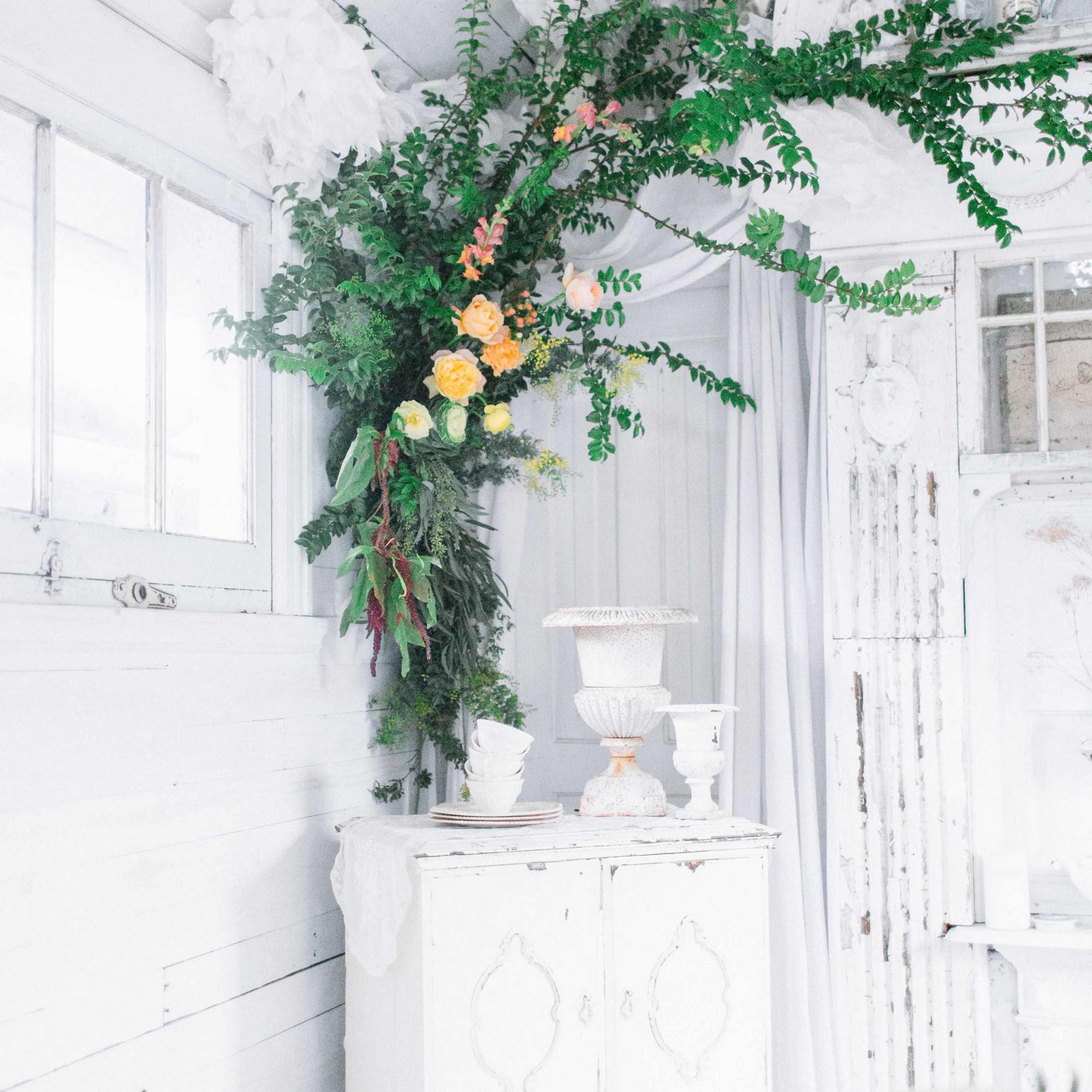 A white wooden cabinet stands in a room adorned with greenery and flowers, complemented by light-colored curtains and rustic decor.