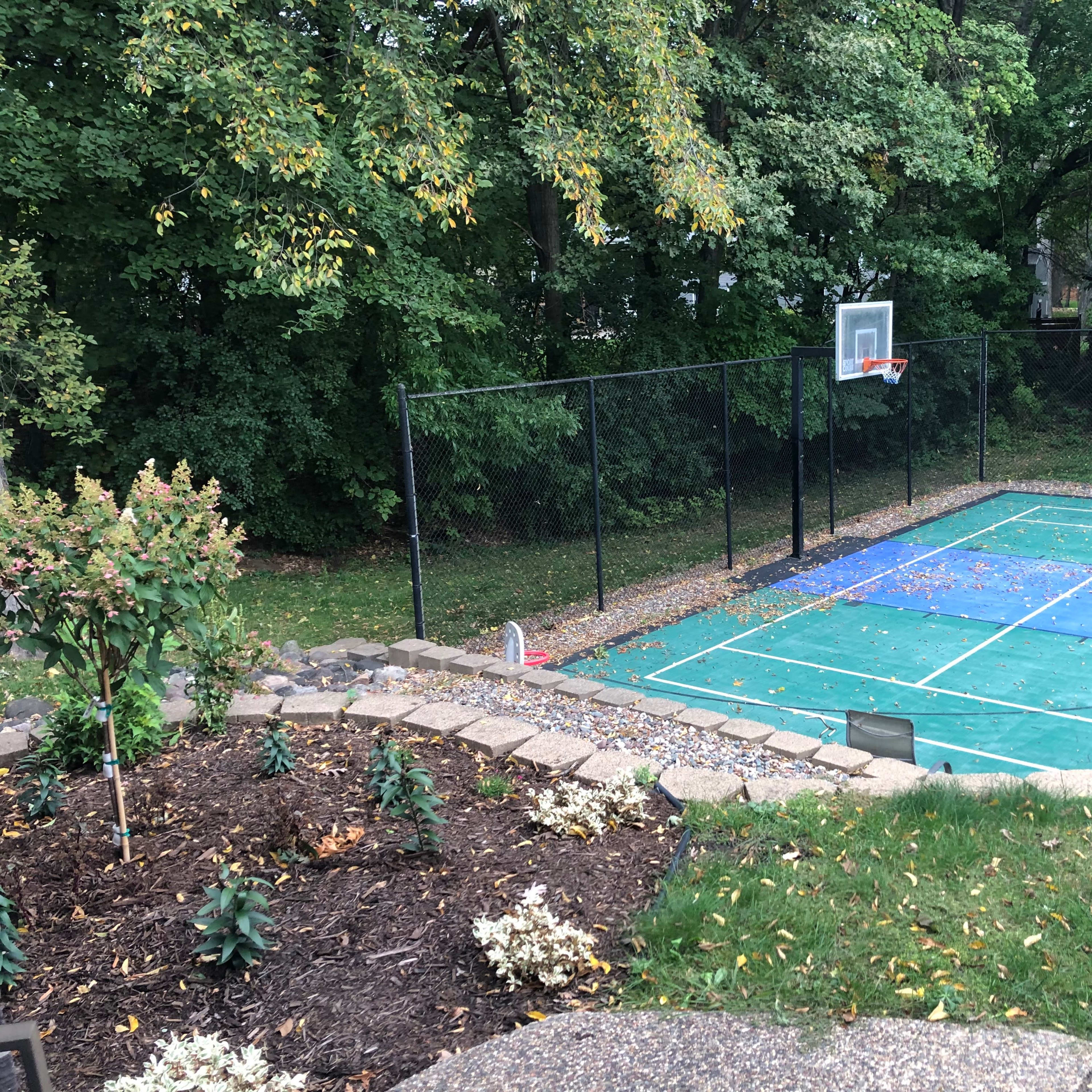 A basketball court surrounded by a chain-link fence, with a garden and lawn area in the foreground.