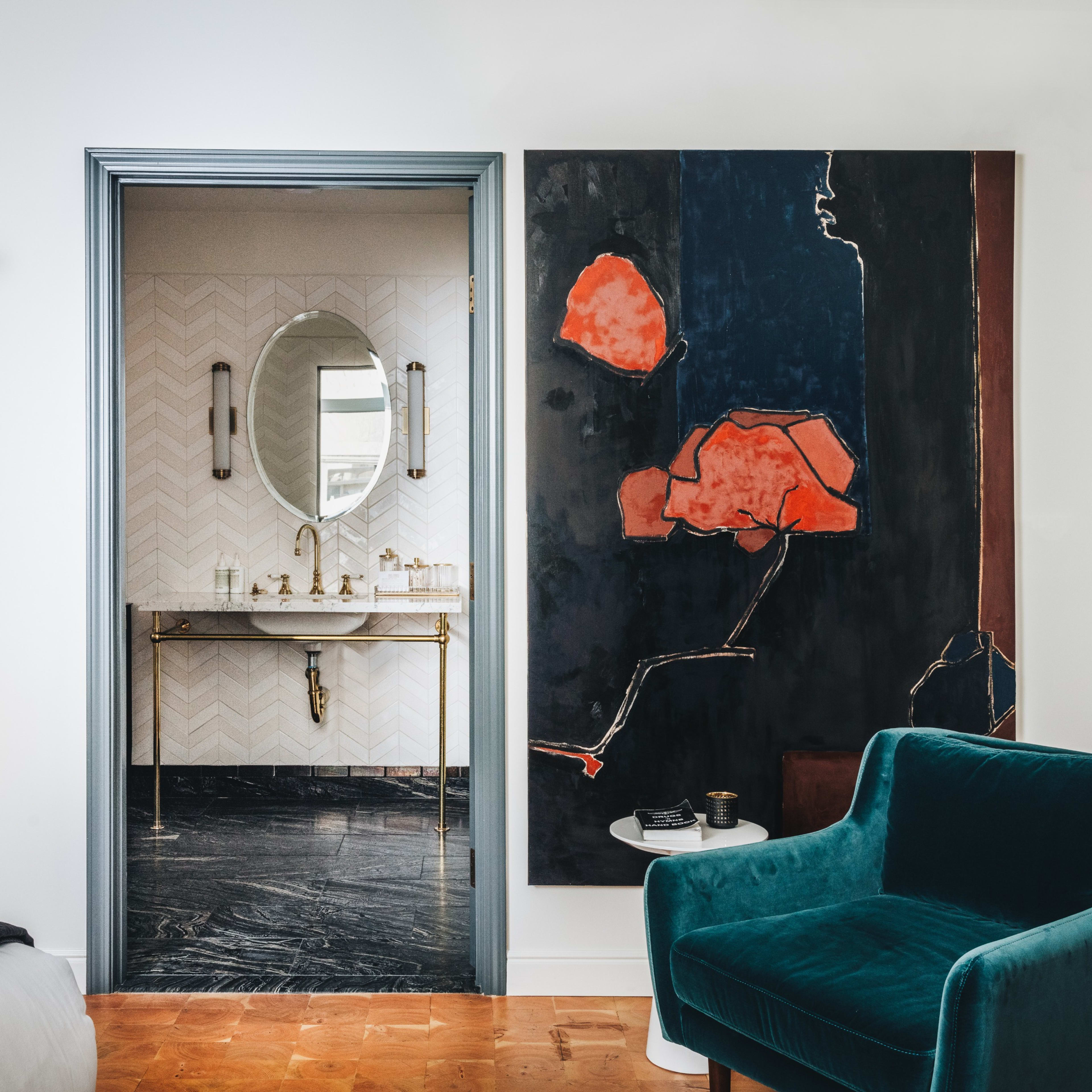 A modern hotel room with a bathroom entrance, featuring a circular mirror, a large abstract painting, and a green armchair next to a small side table.