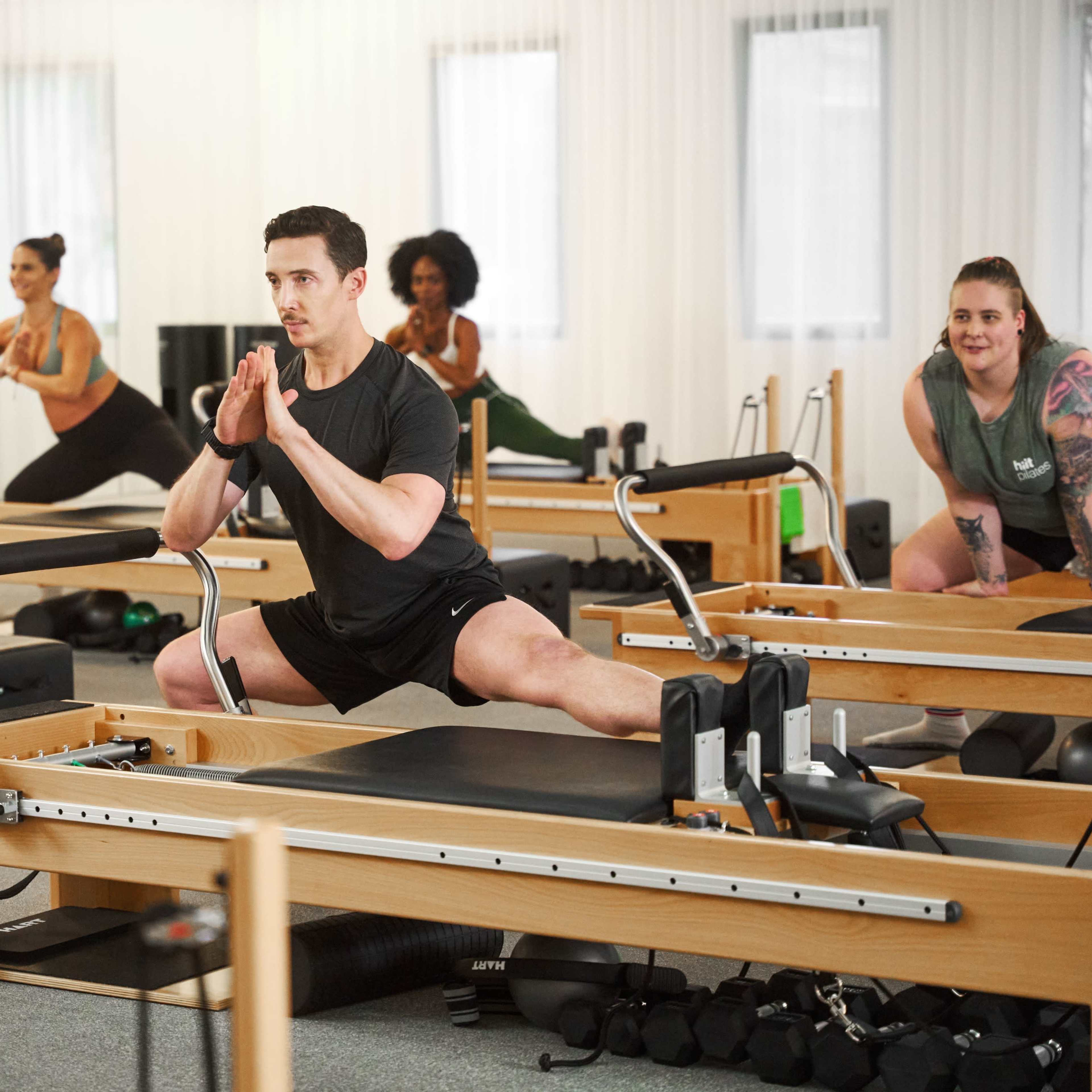A group of people is participating in a Pilates class, using reformer machines in a well-lit studio.