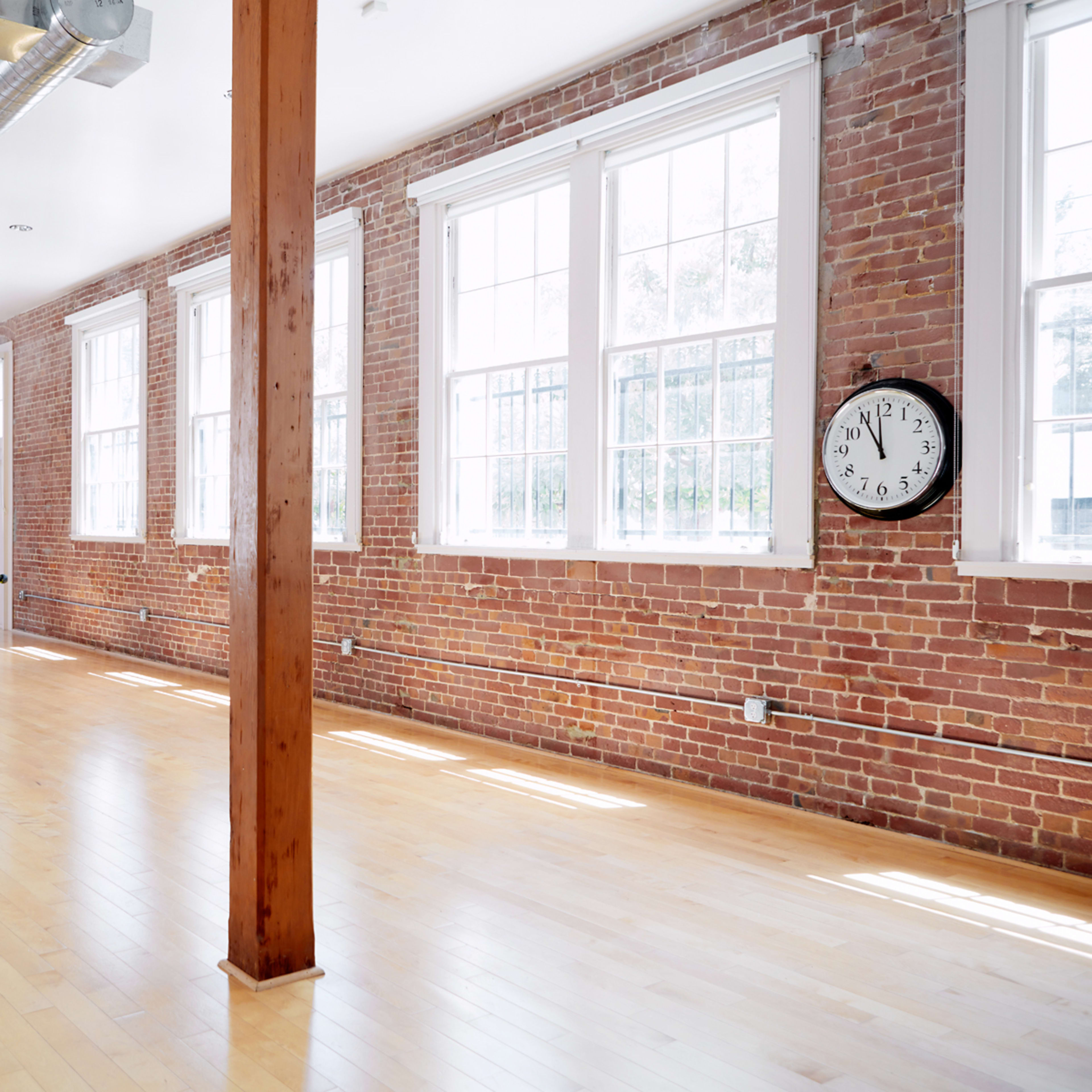 An empty room with hardwood floors, exposed brick walls, large windows, and a clock mounted on the wall.