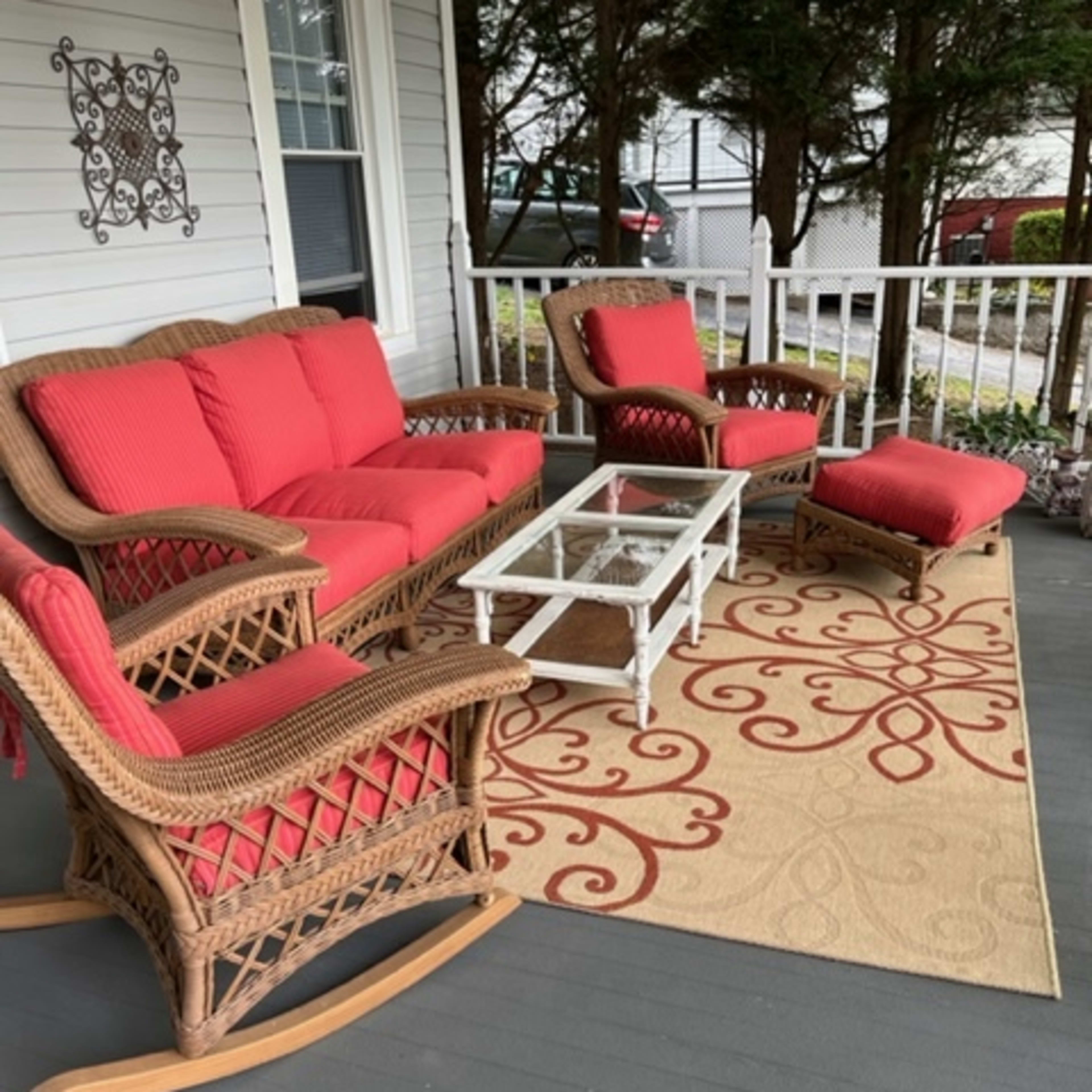 A porch features a wicker seating arrangement with red cushions, a glass coffee table, and a decorative area rug.