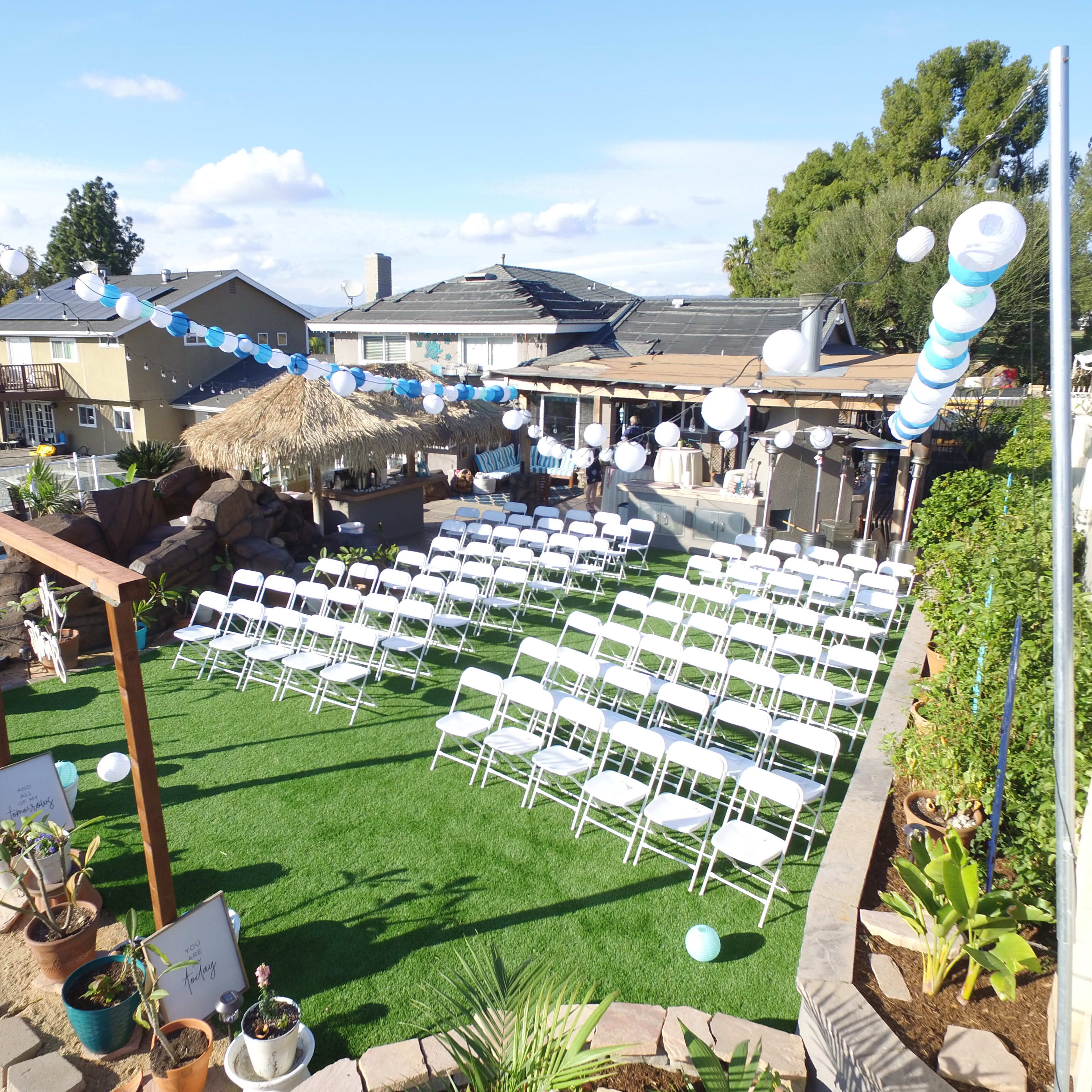 A neatly arranged outdoor seating area with white chairs set up on green grass, surrounded by decorative balloons and a thatched-roof structure.