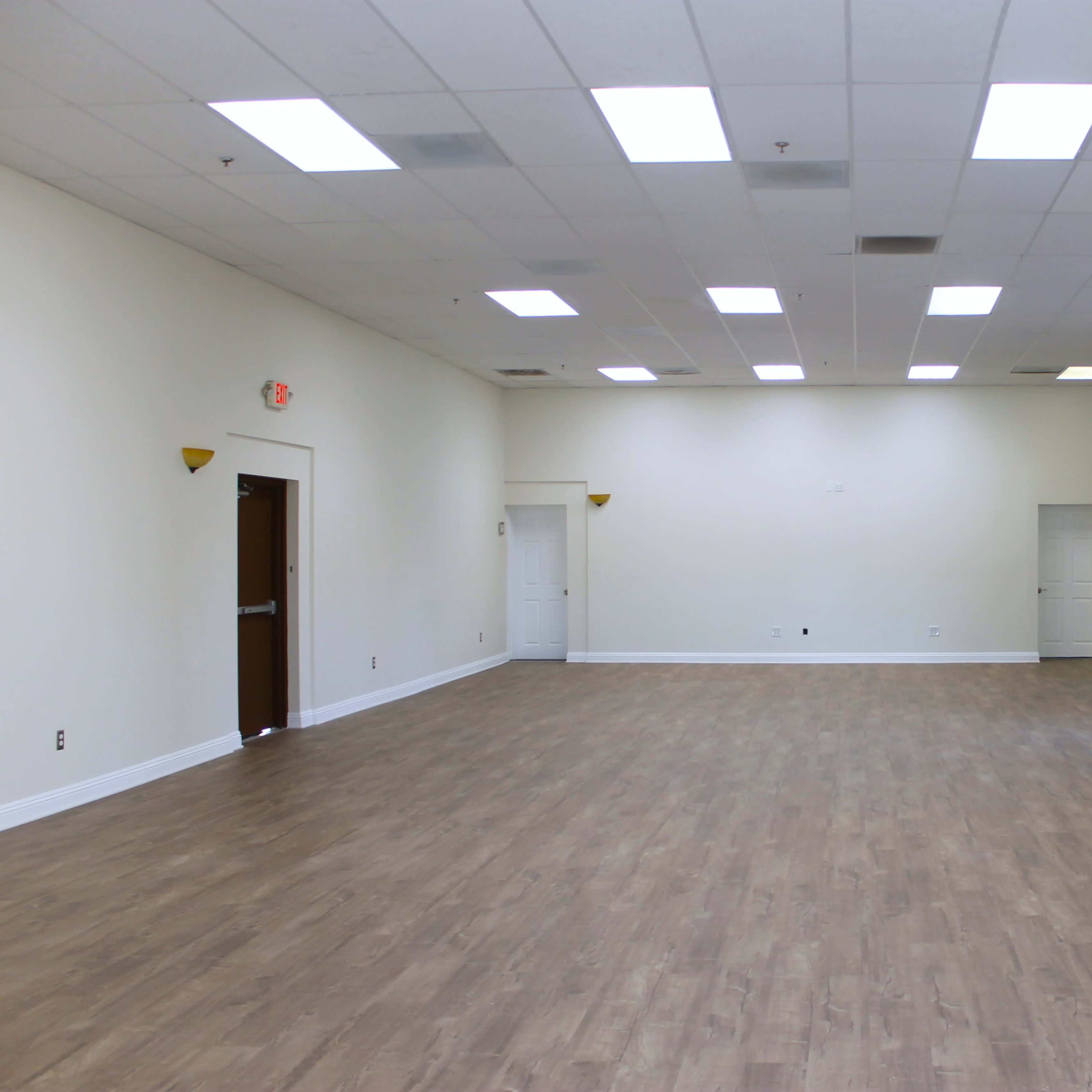 A spacious, empty room with a wooden floor, white walls, and multiple doors along the perimeter.