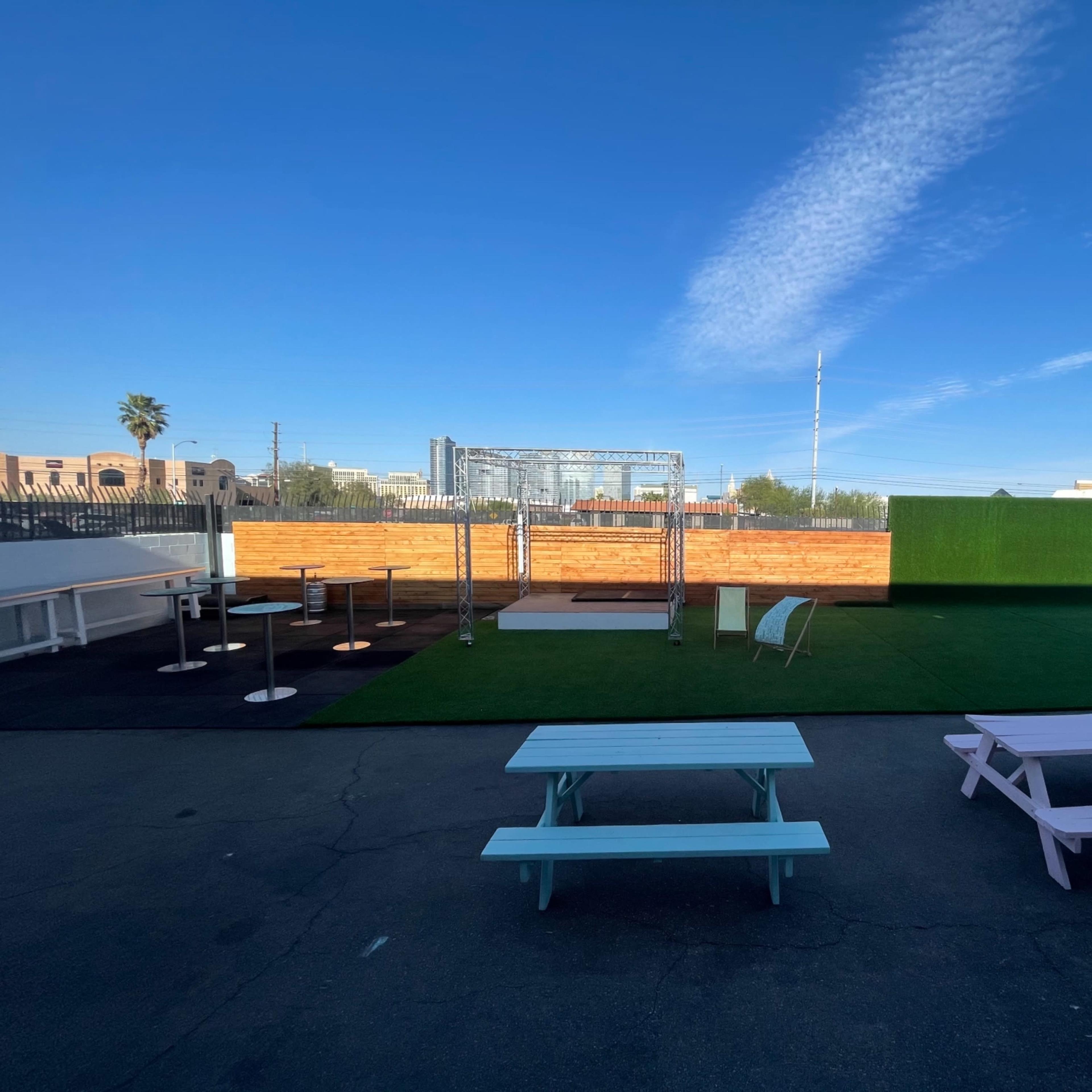 An outdoor venue with a green turf area, picnic tables, a wooden stage, and a backdrop of a blue sky.