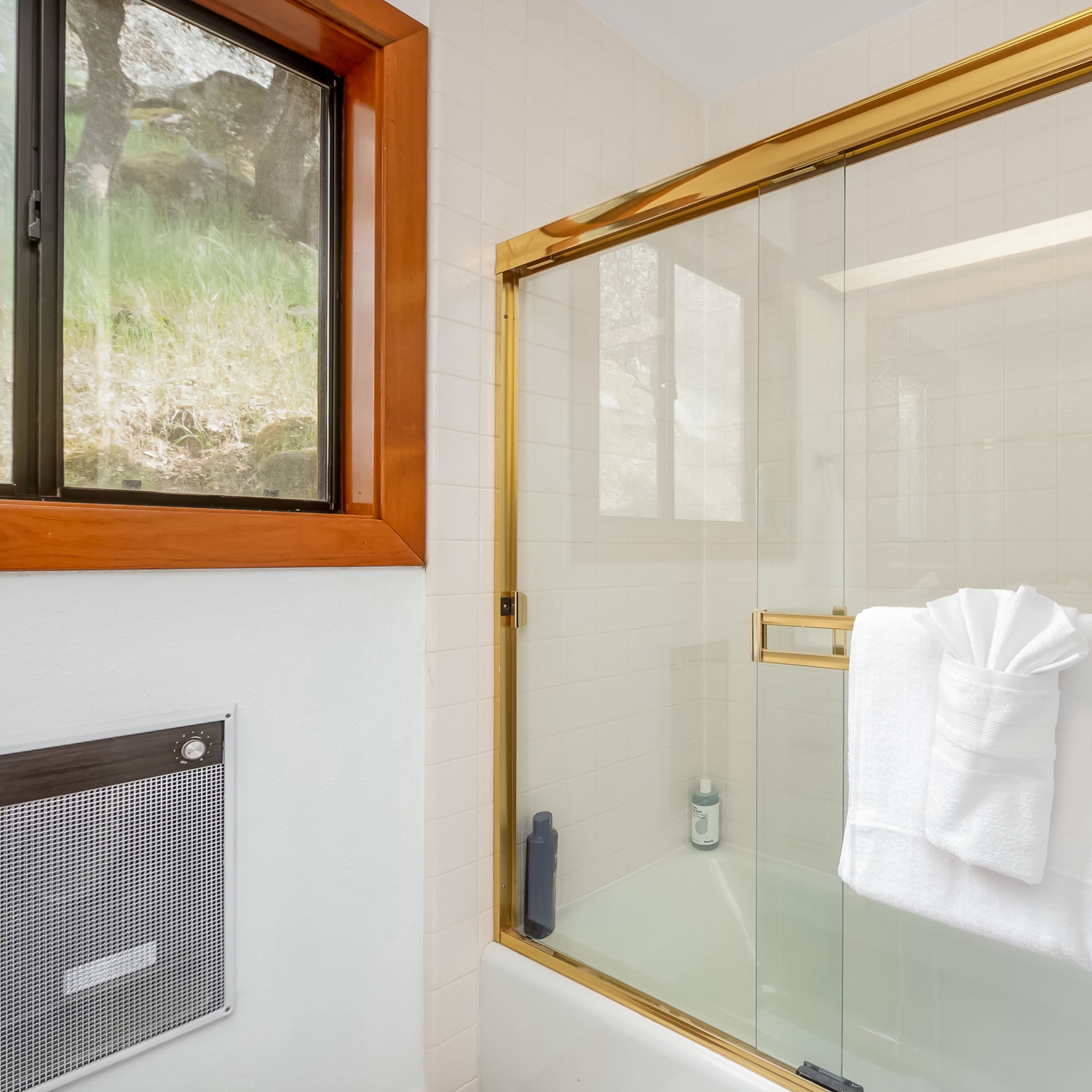 A bathroom with a glass shower enclosure, a white towel neatly folded on the edge of a bathtub, and a window allowing natural light to enter.