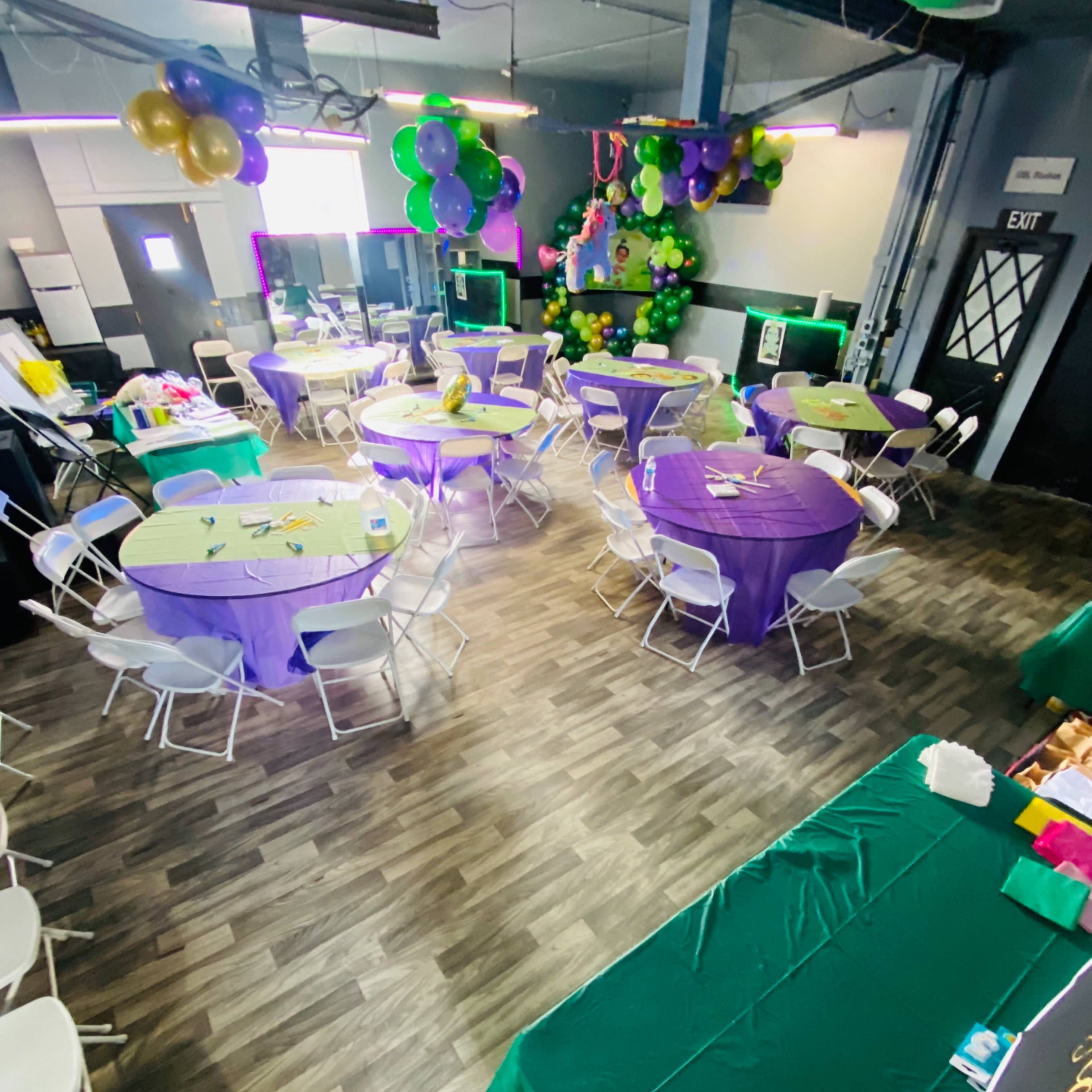 A decorated event space with round tables covered in purple and green tablecloths, surrounded by balloons and a festive atmosphere.