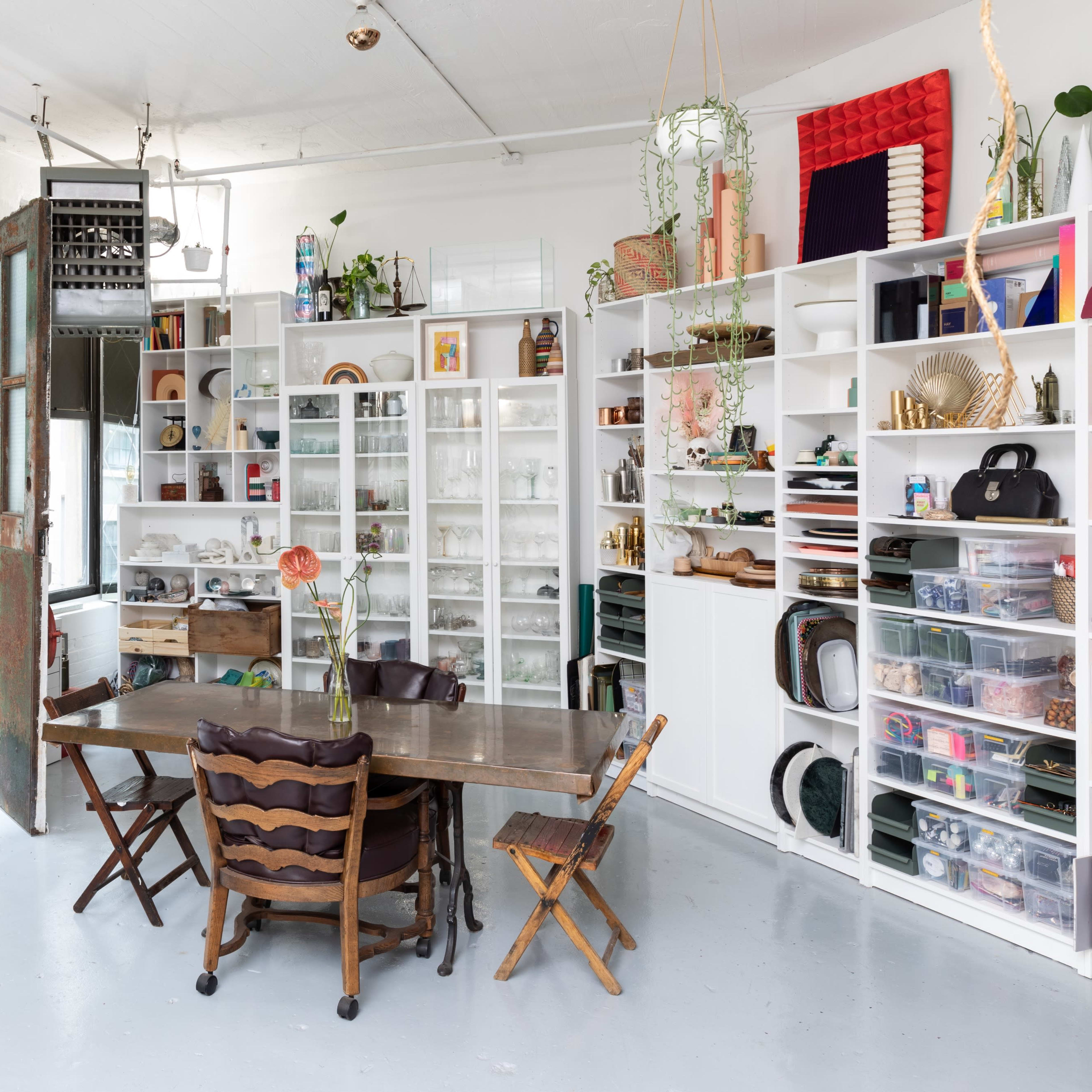 A spacious room with large windows, a wooden table surrounded by chairs, and a shelving unit filled with various items.