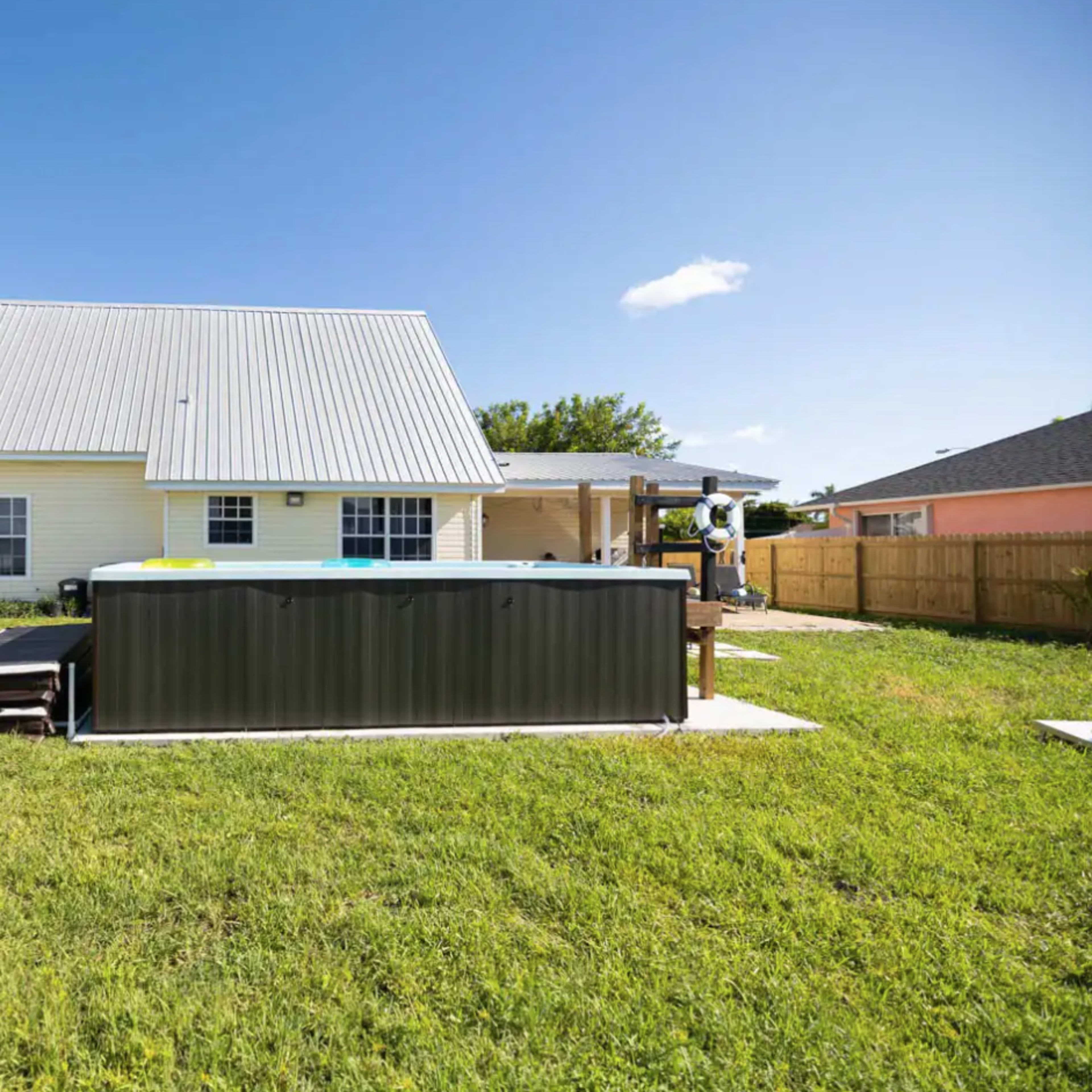 A single-story house with a metal roof and a hot tub situated in a grassy backyard surrounded by a wooden fence.