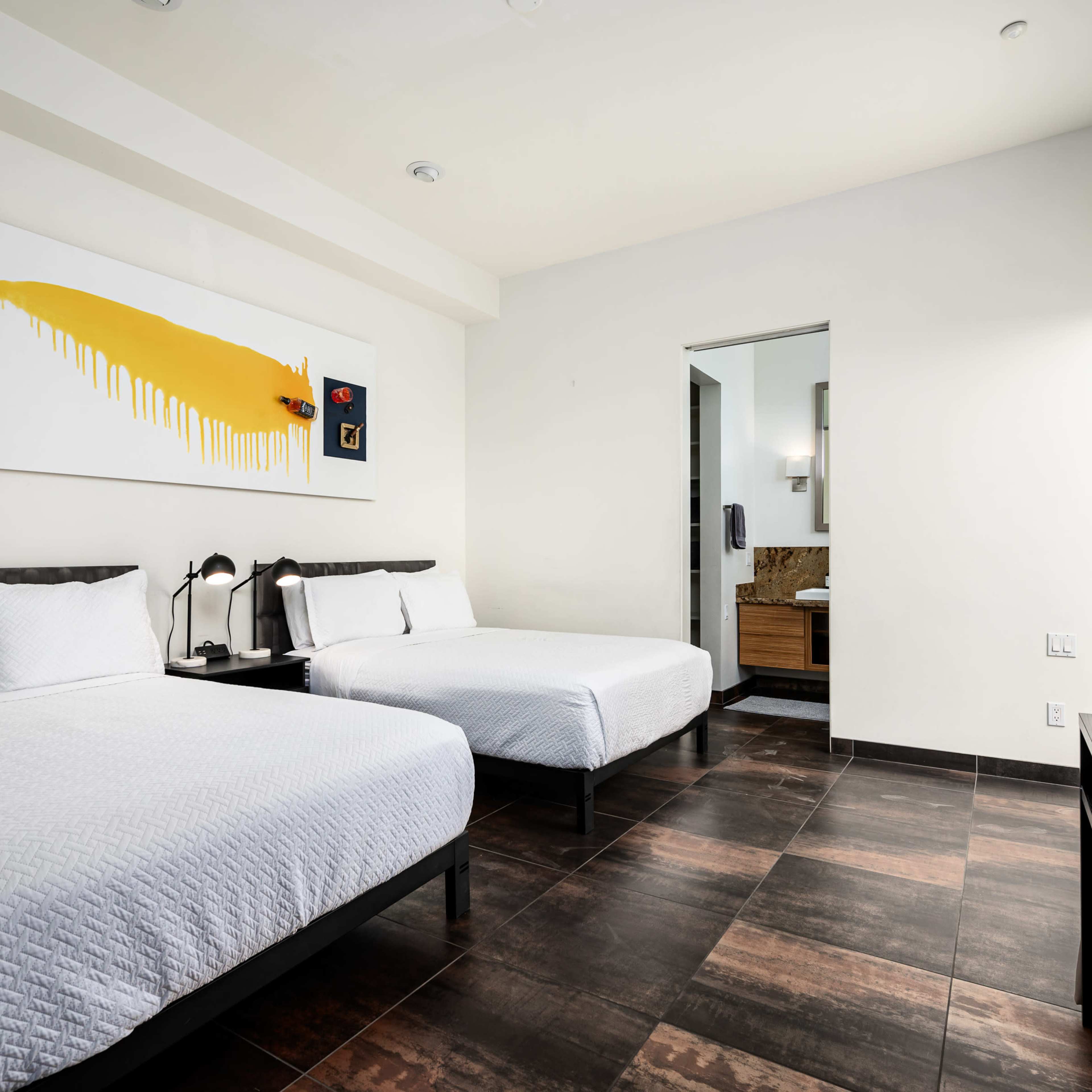 A hotel room with two beds, a large piece of abstract art on the wall, and an adjacent bathroom visible in the background.