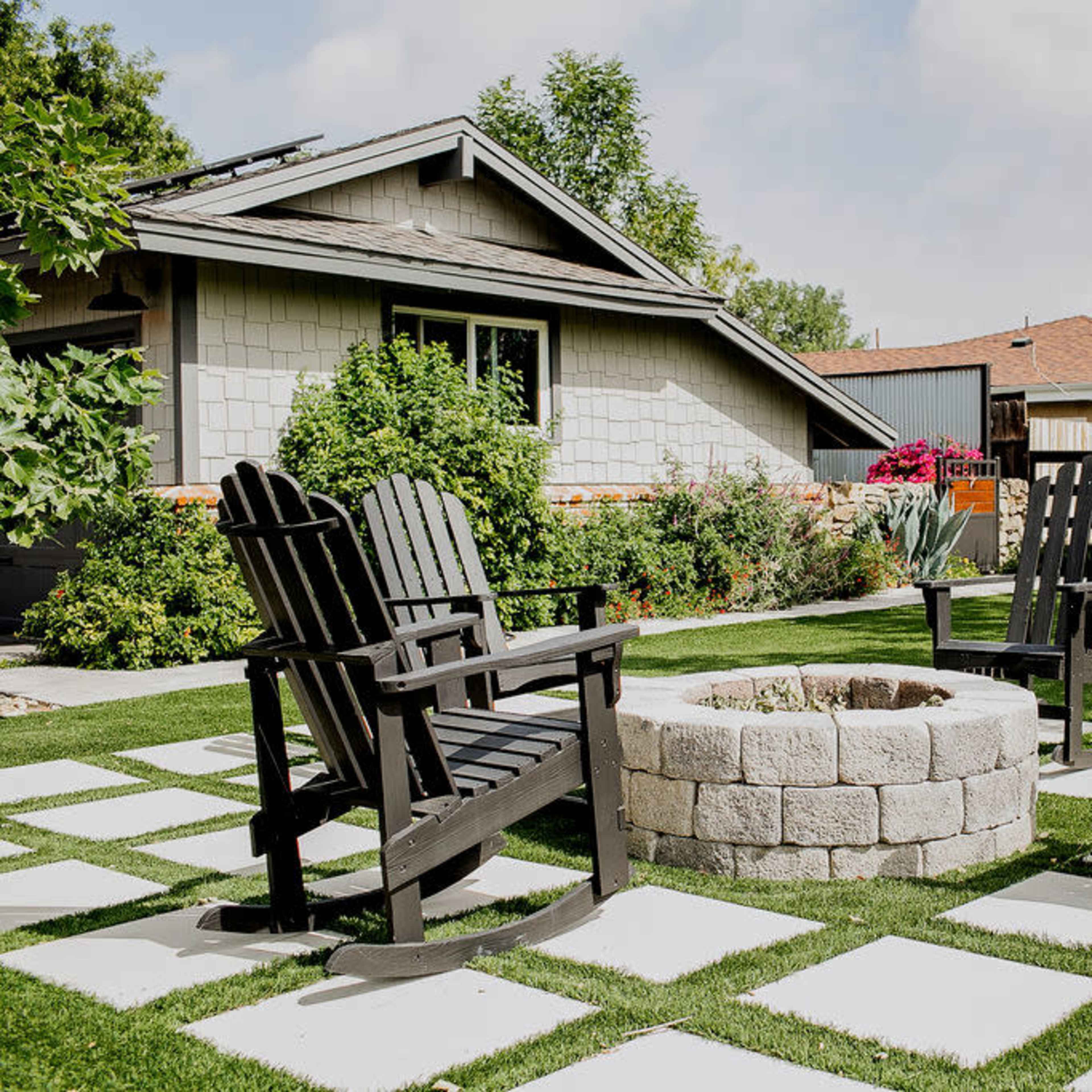 Four black rocking chairs are arranged around a stone fire pit on a grassy area with paved stone paths and a house in the background.