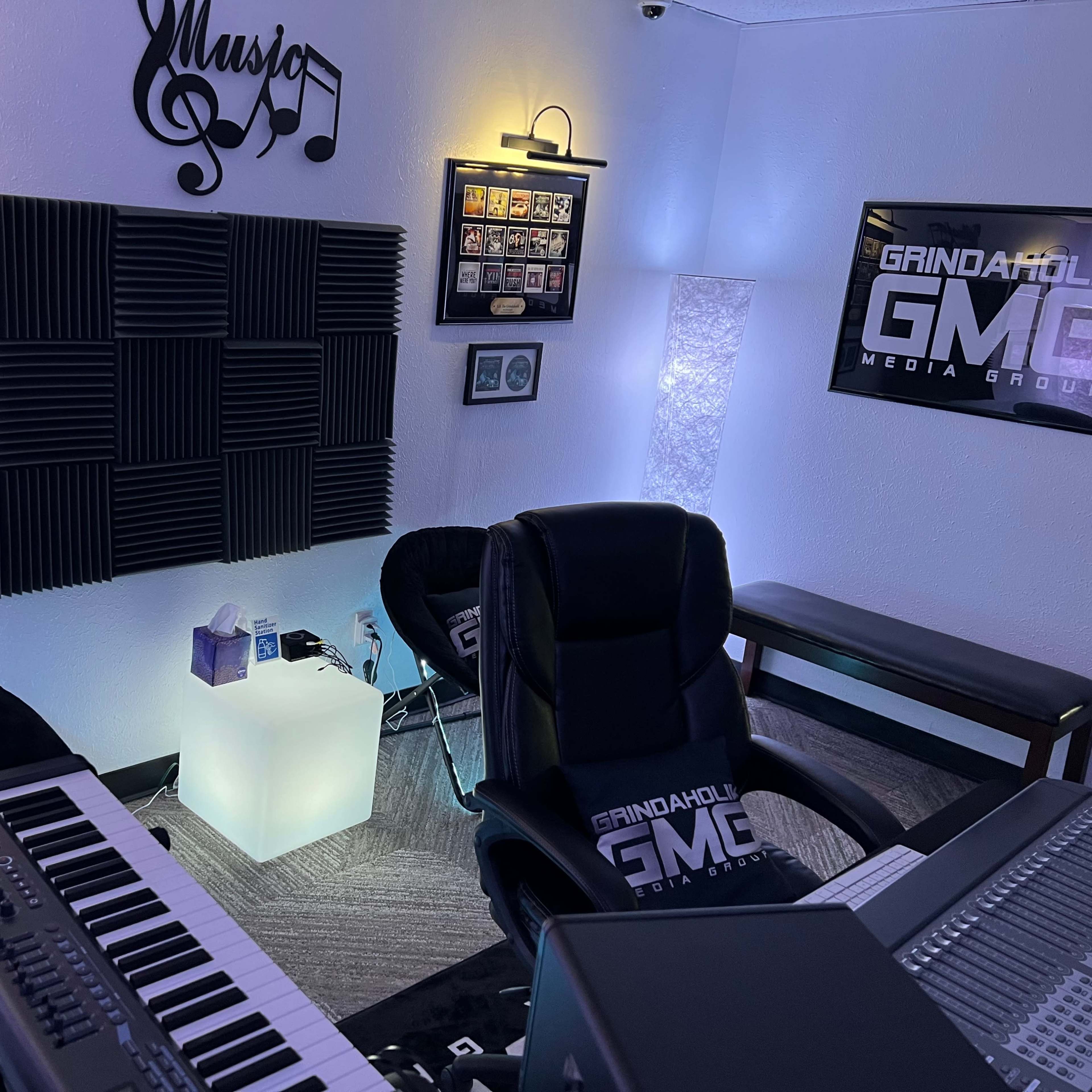 A music studio featuring a keyboard, black chairs, acoustic panels on the walls, and framed music awards.