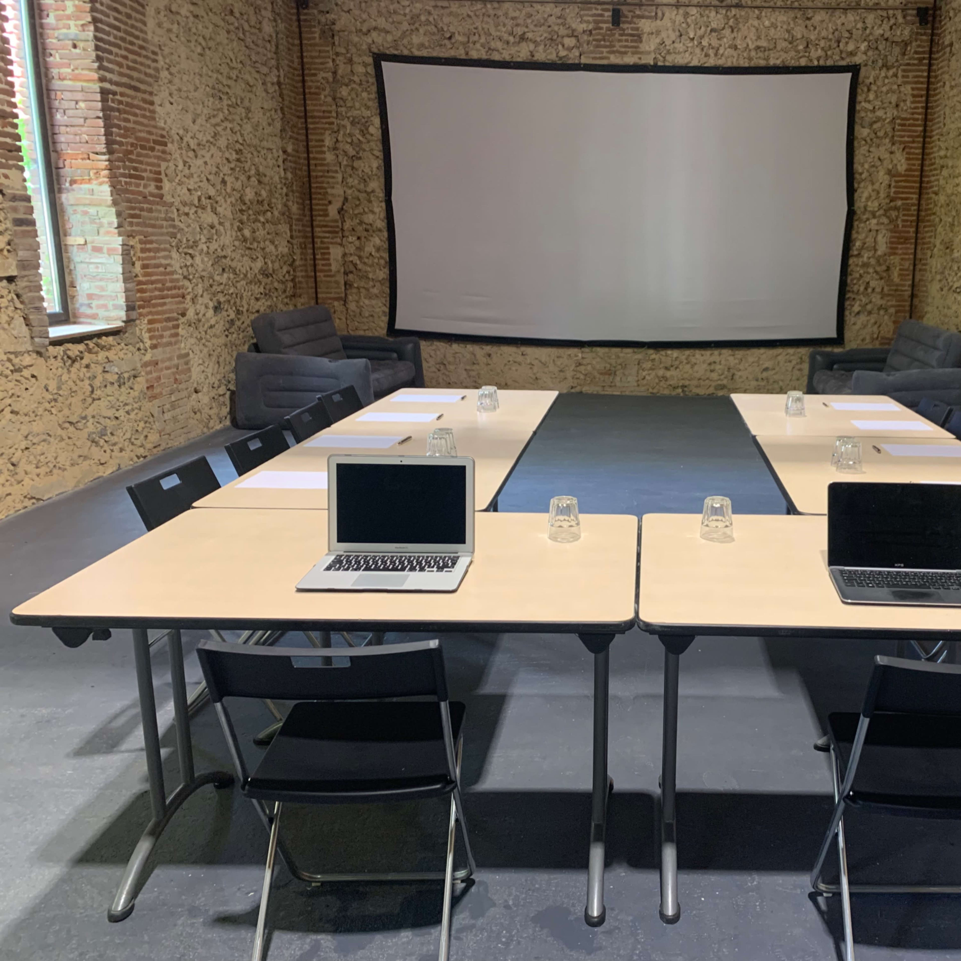 A conference room with two long tables set up in a U-shape, each equipped with laptops and glass containers, facing a large projection screen.