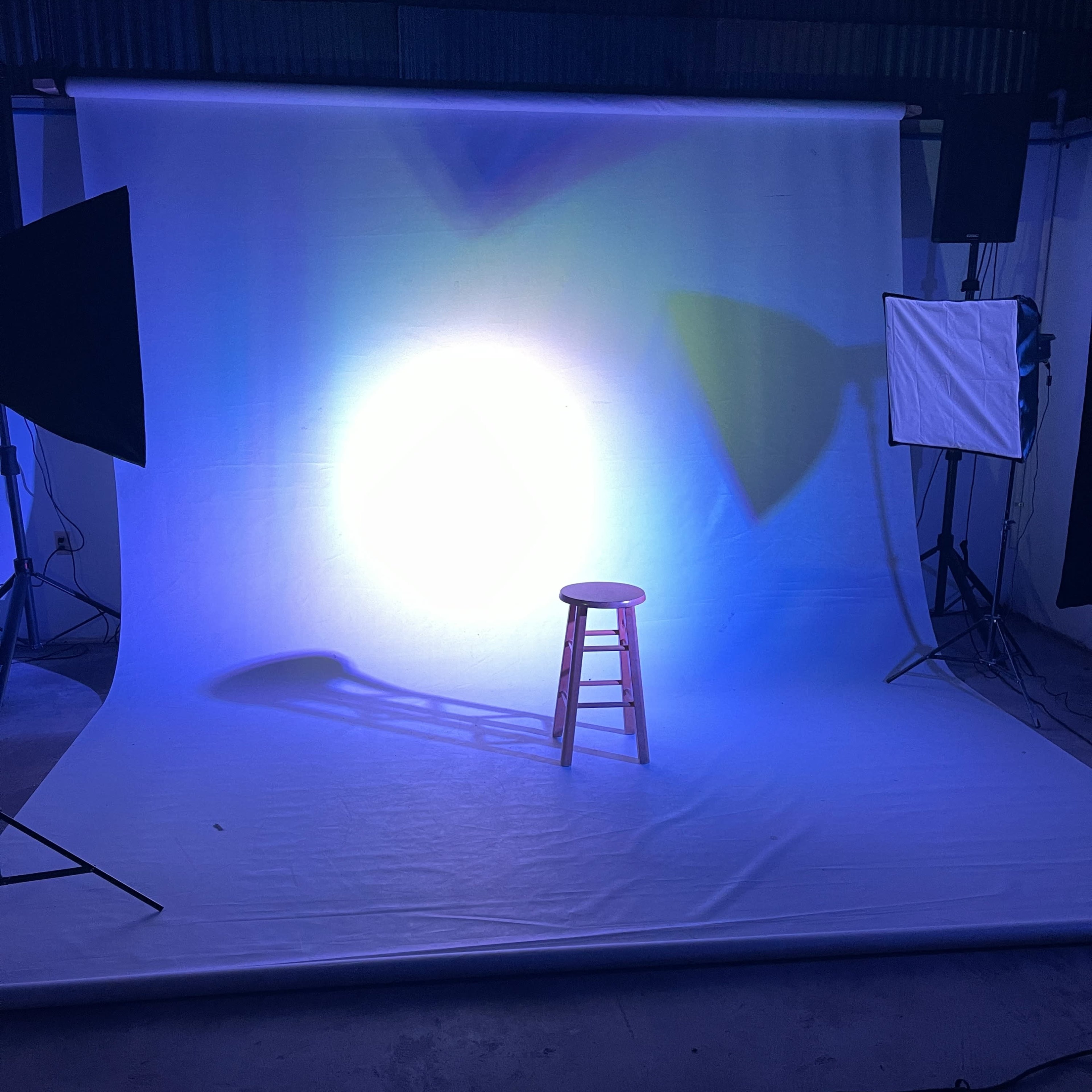 A brightly lit photography set with a wooden stool positioned on a white backdrop, surrounded by studio lighting equipment.