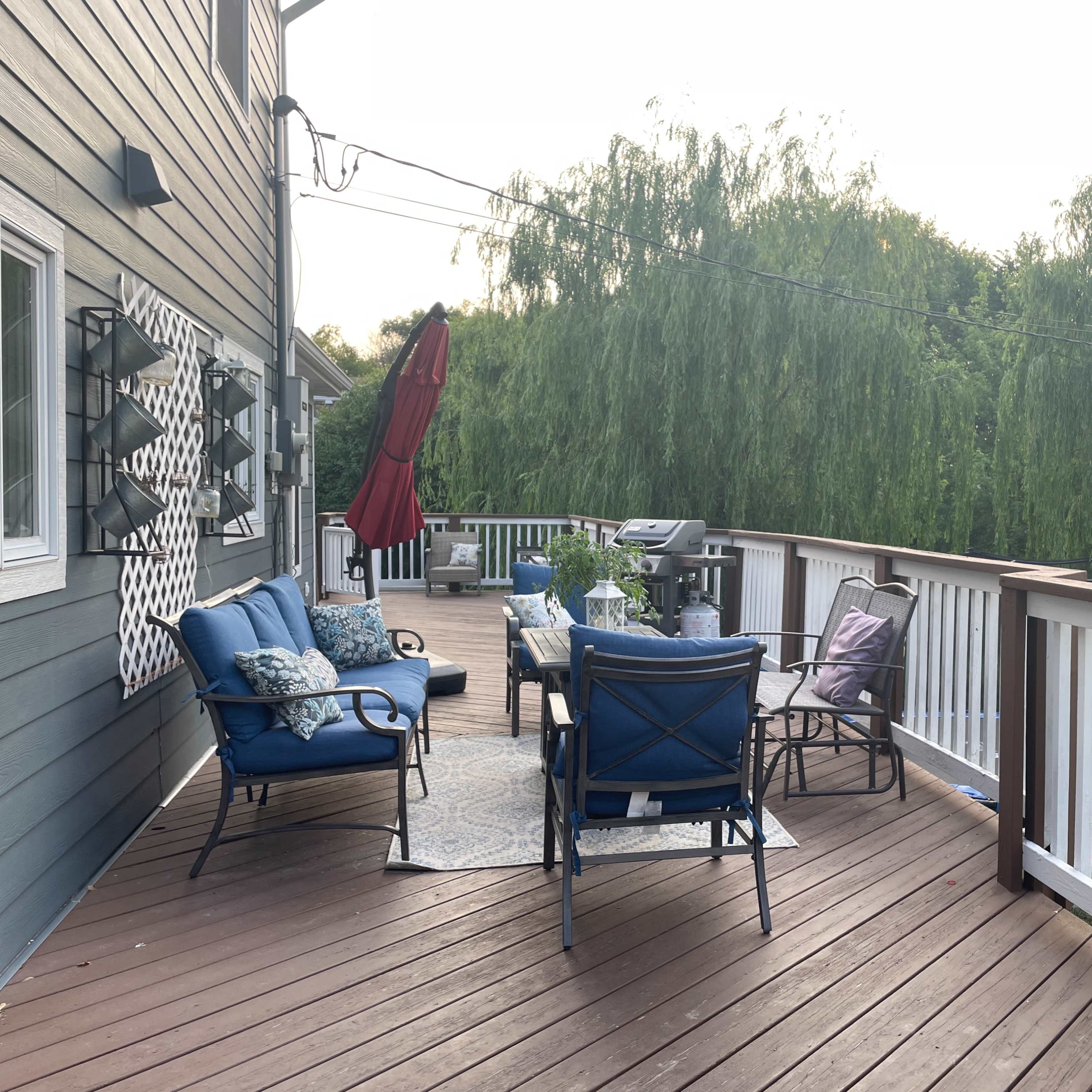 A wooden deck with blue seating, a table, and a red umbrella, surrounded by greenery.