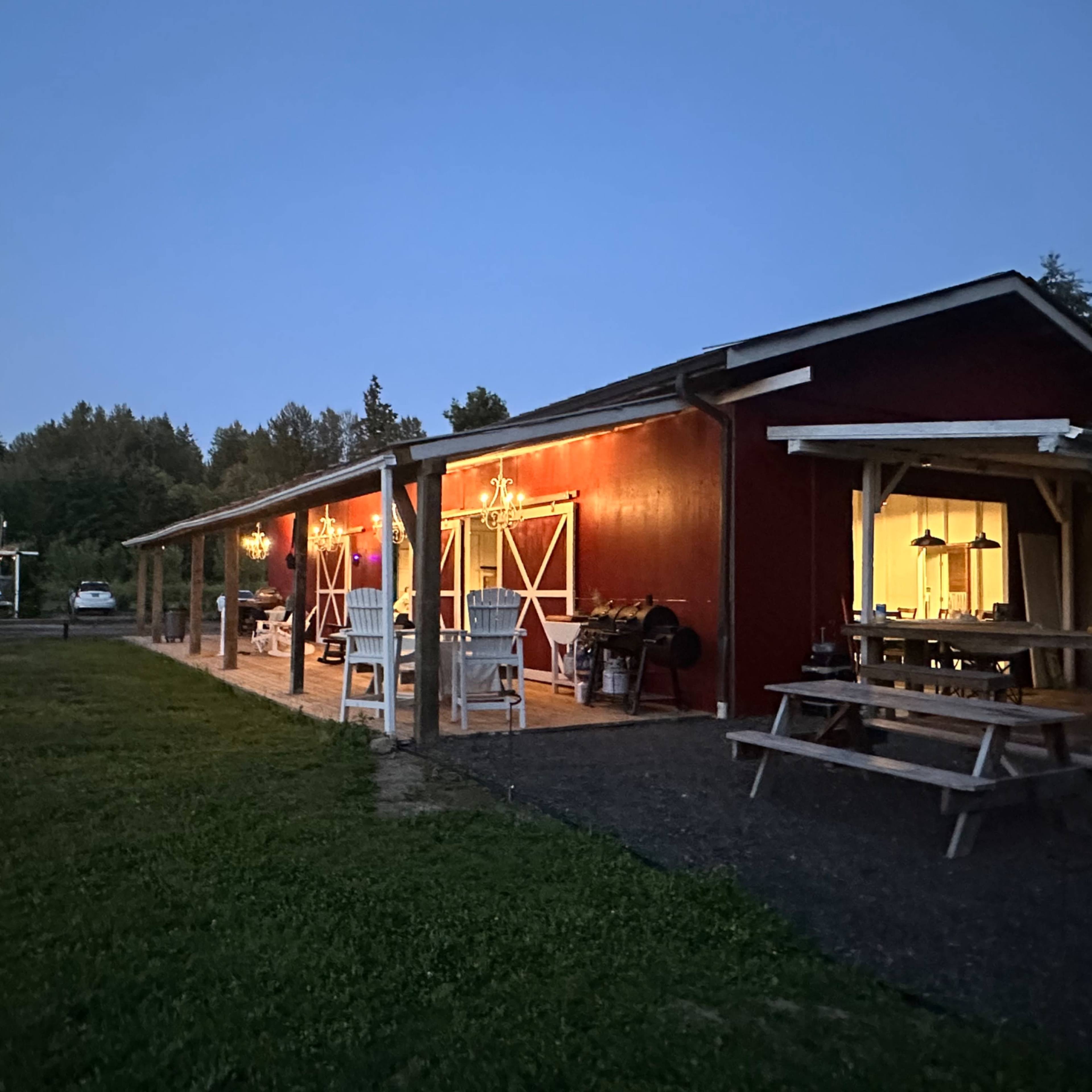 A red wooden building with outdoor seating and lights, surrounded by grass and trees, under a twilight sky.