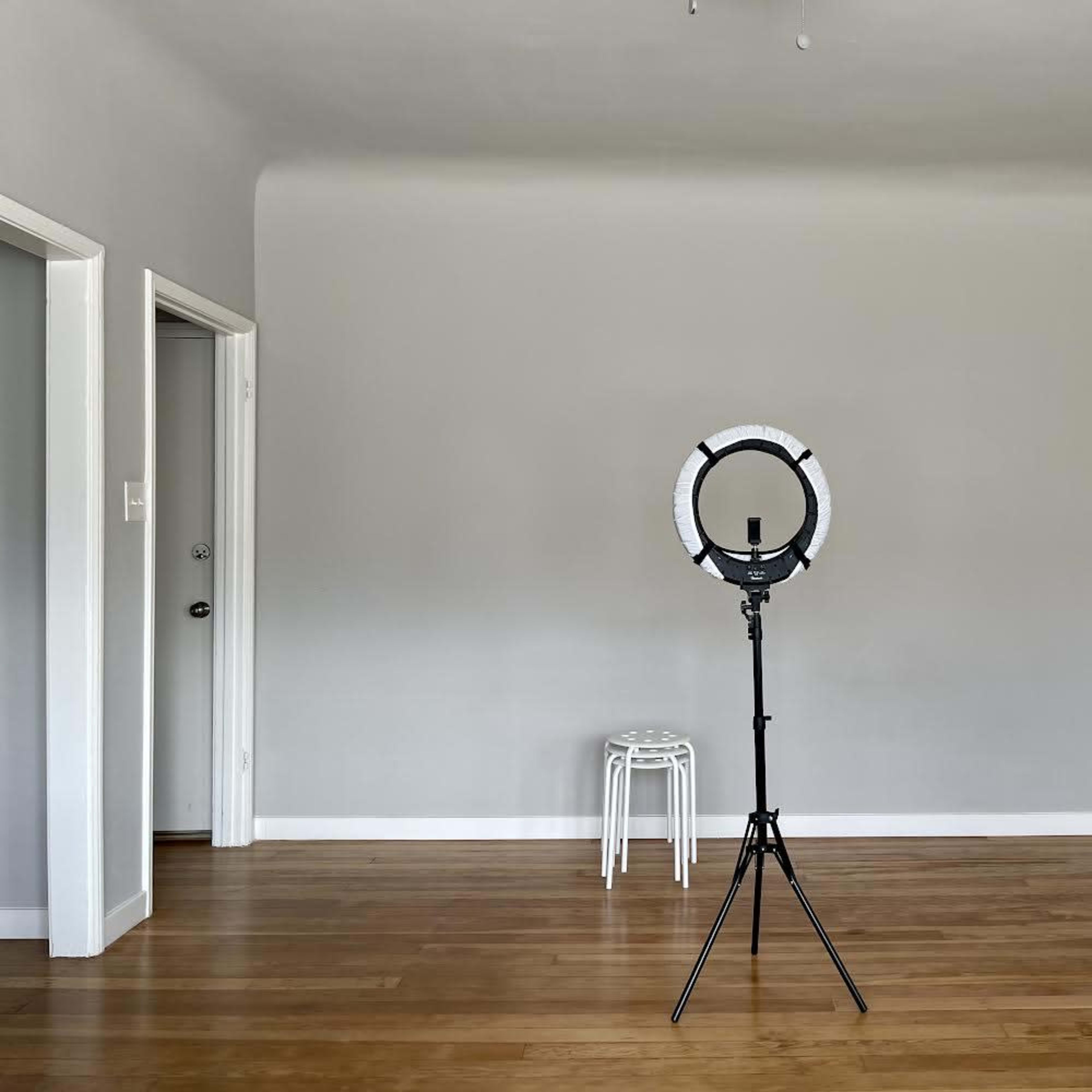 A ring light on a tripod stands in the center of an empty room with light wooden floors and gray walls.