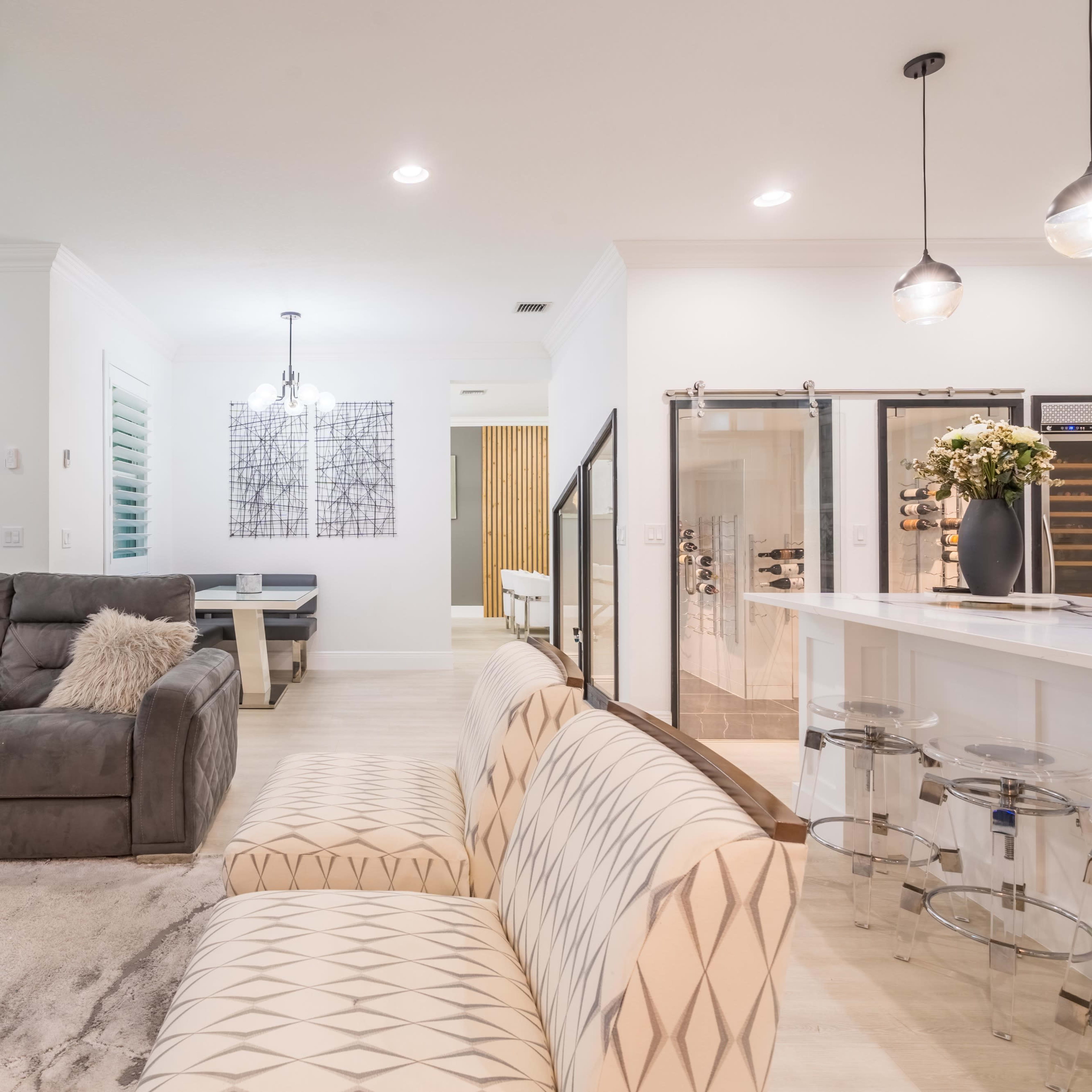 A modern living space featuring a gray sectional sofa, a patterned white couch, and a kitchen with a bar and dining area.