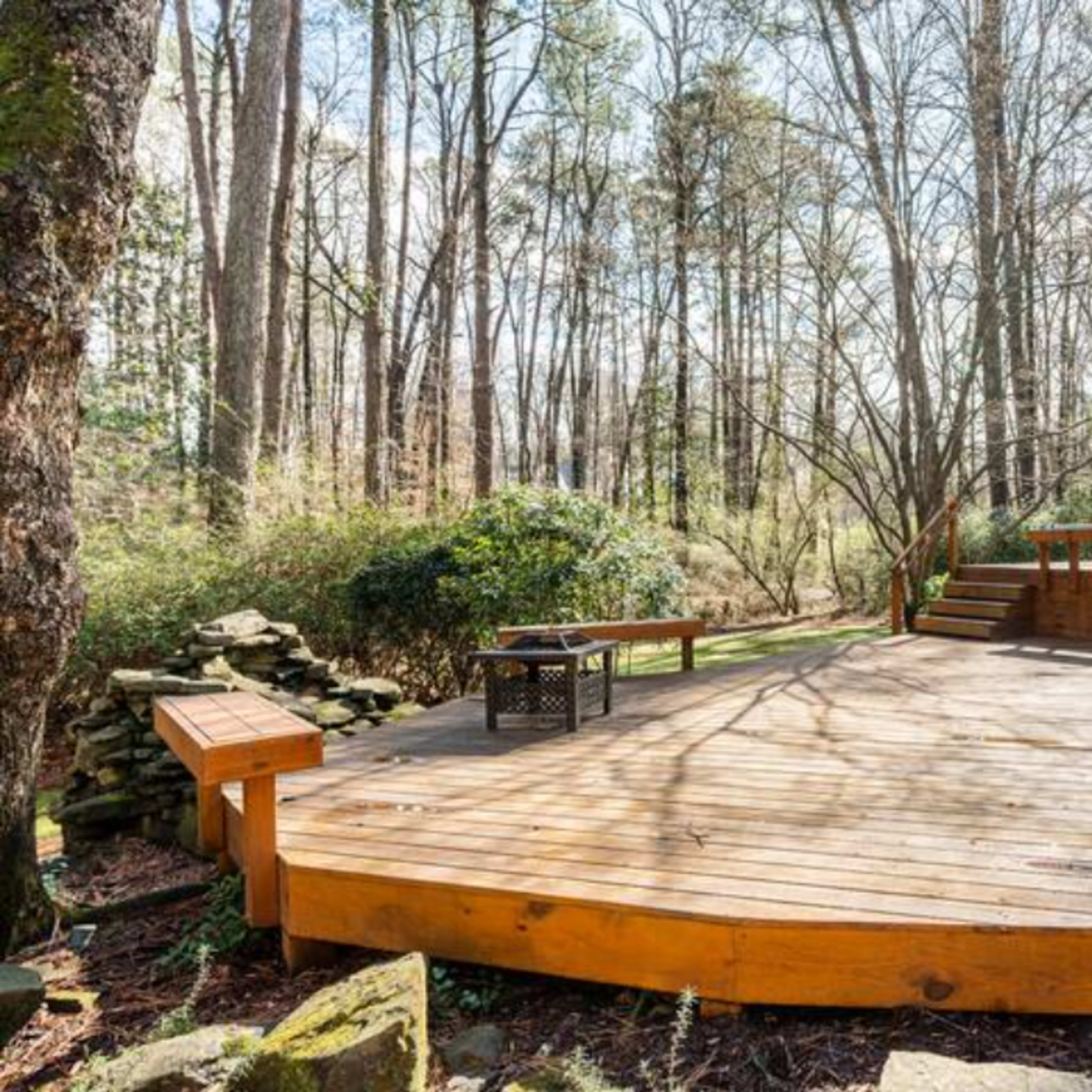 A wooden deck surrounded by trees and greenery features a seating area and a grill setup.