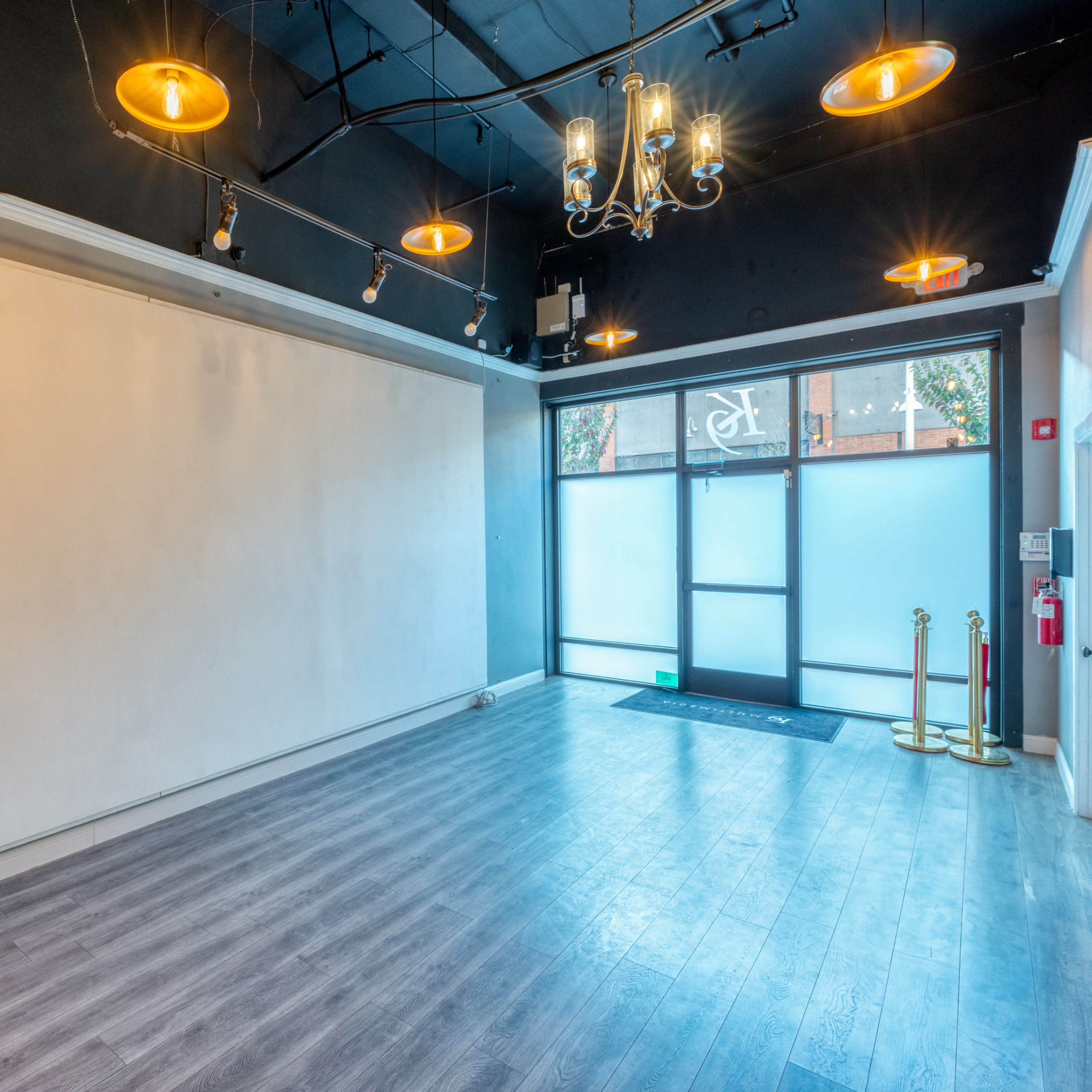 A vacant commercial space with a large, blank wall, hardwood flooring, and a glass entrance door illuminated by pendant lights.