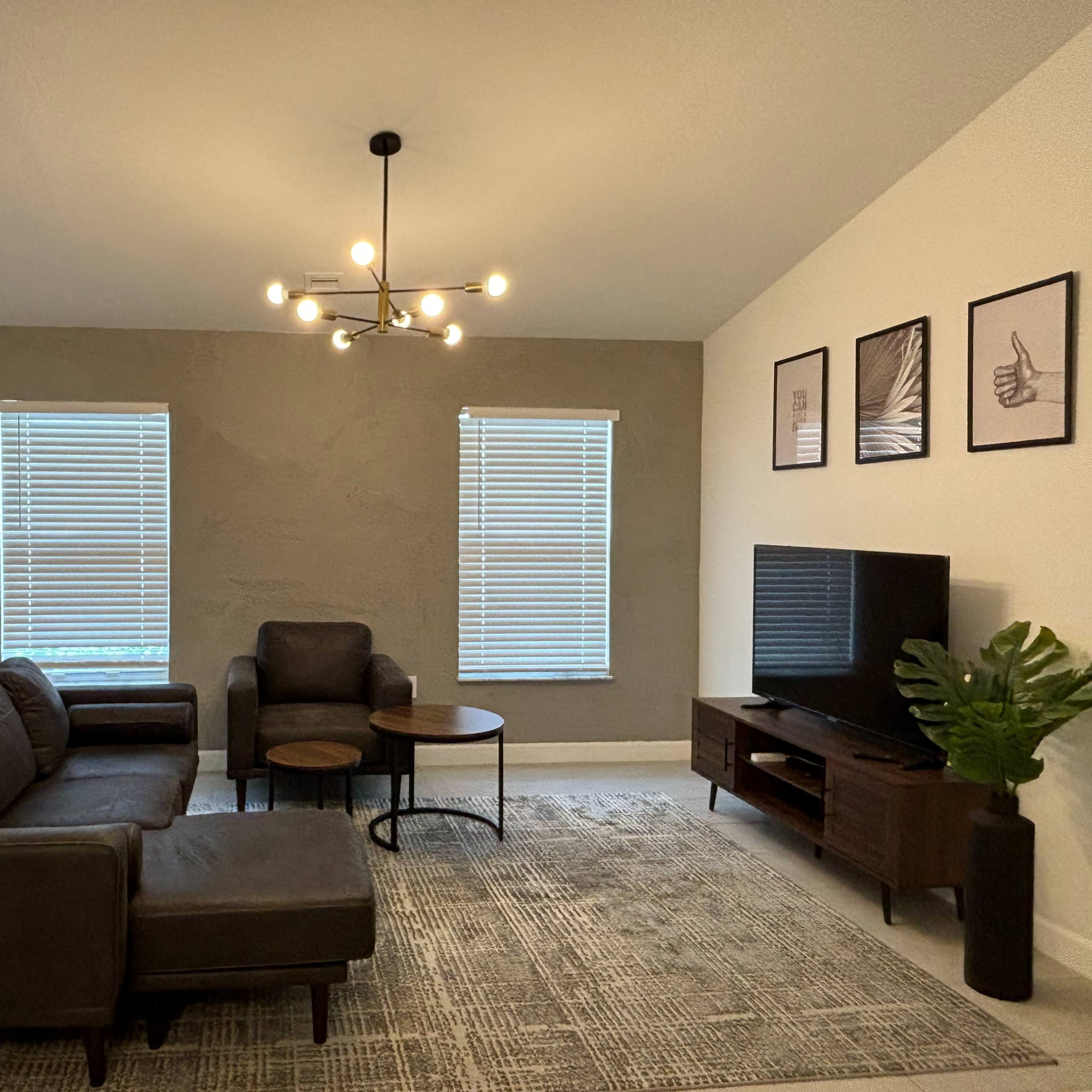 A living room features a sectional sofa, two windows with blinds, a television on a stand, and a round coffee table with a plant nearby.
