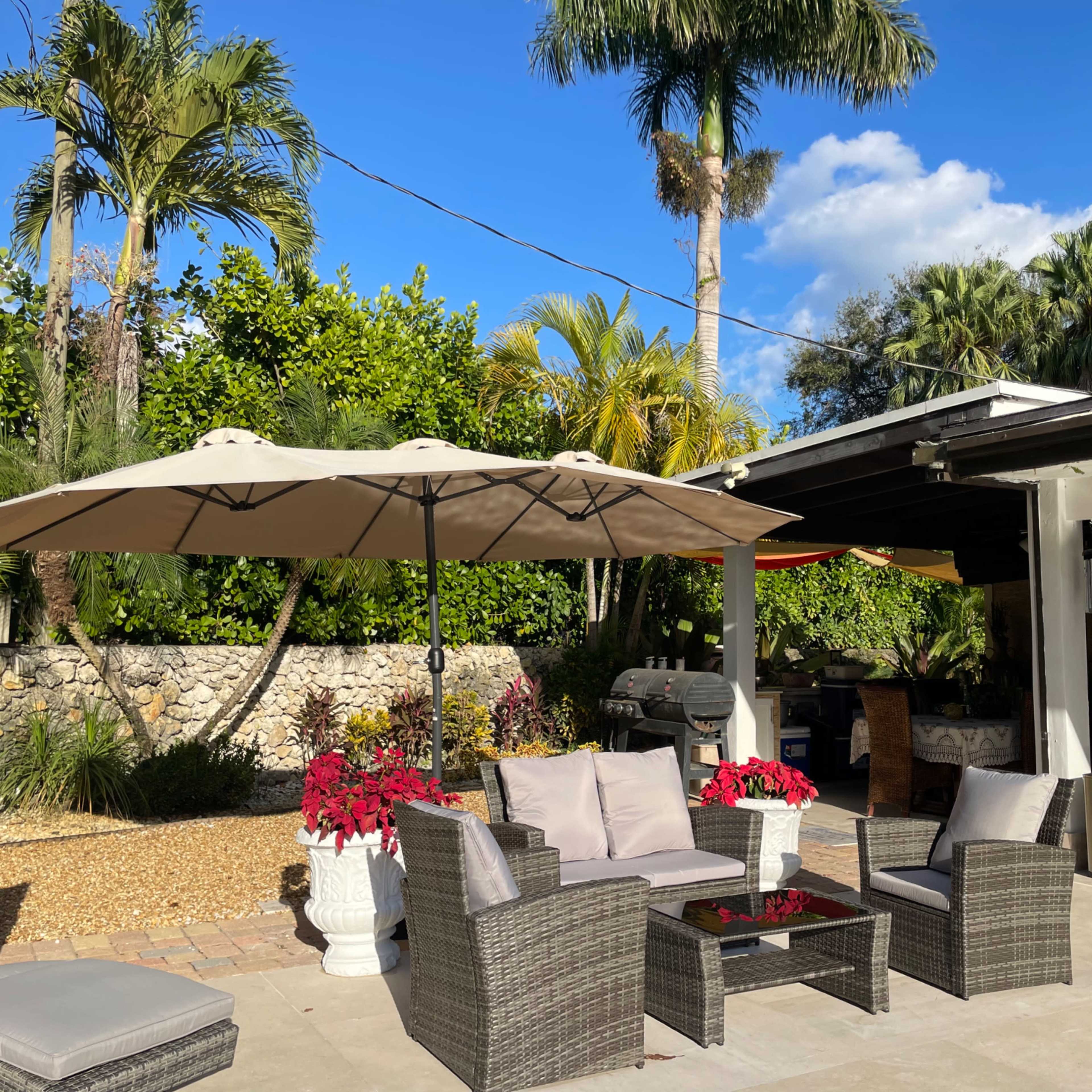 A backyard patio with wicker furniture, a large umbrella, and potted plants surrounded by lush greenery.