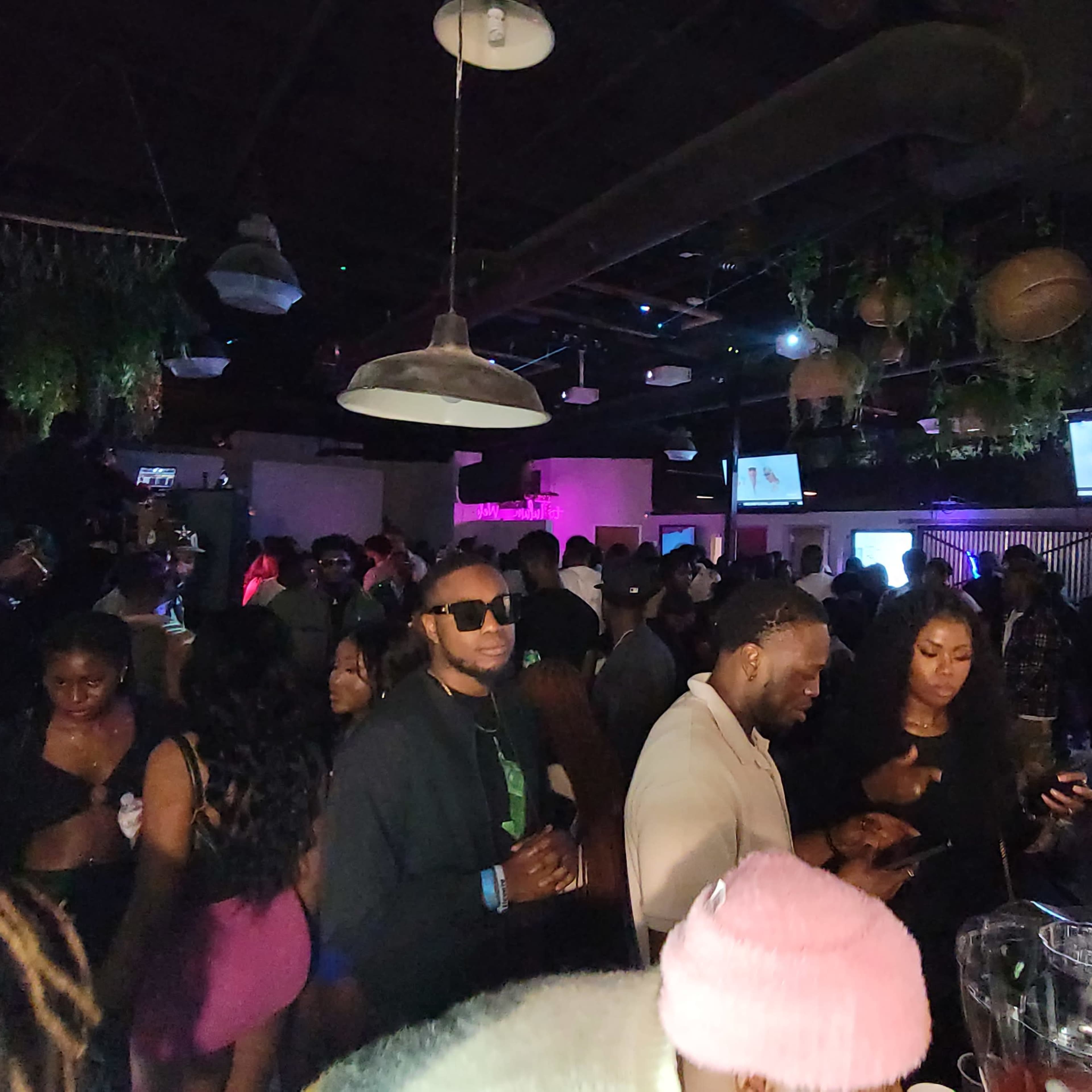 A crowded nightclub atmosphere features patrons socializing and using their phones amidst colorful lighting and screens displaying visuals.