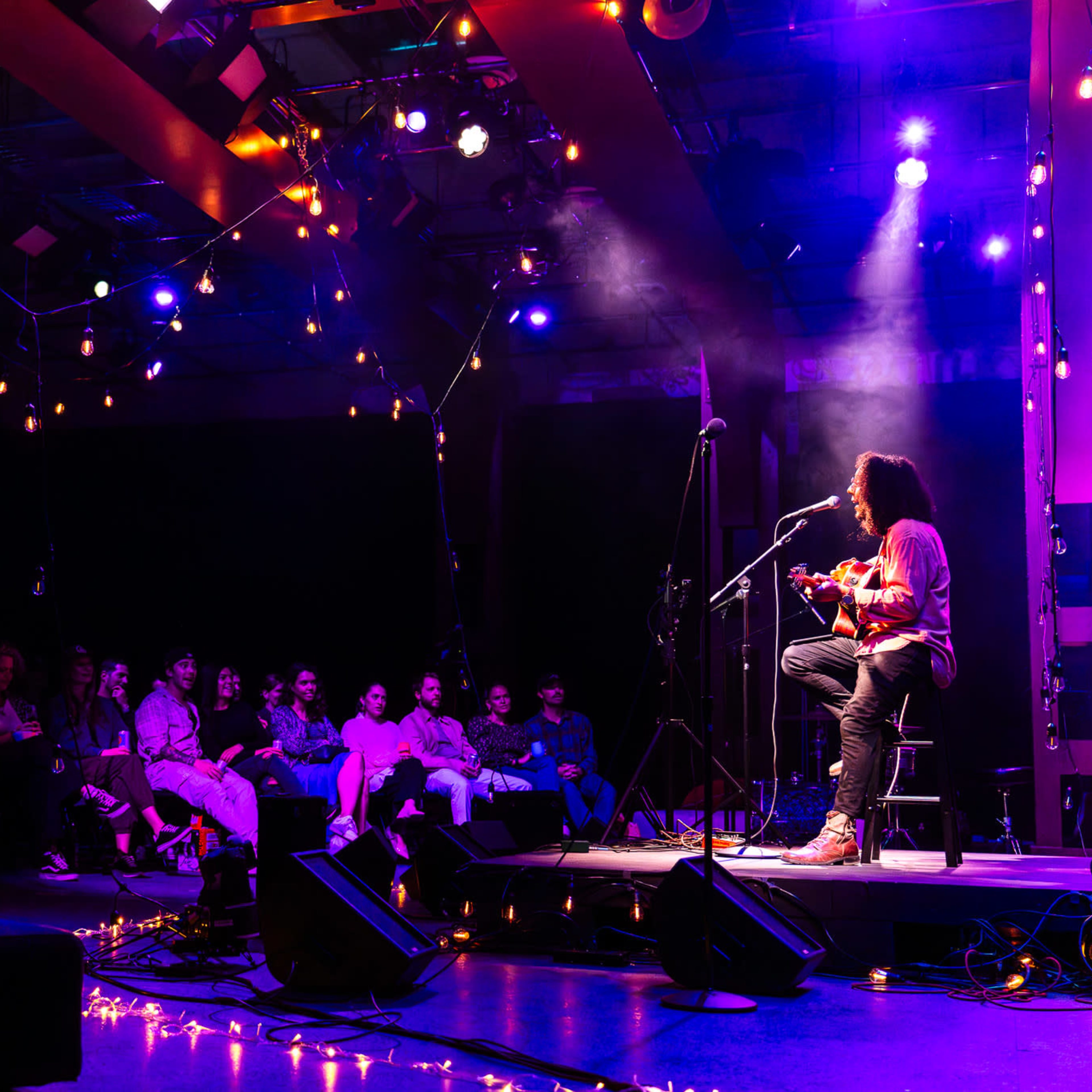 A musician performs on stage with an acoustic guitar in front of an audience seated in a dimly lit venue adorned with string lights.