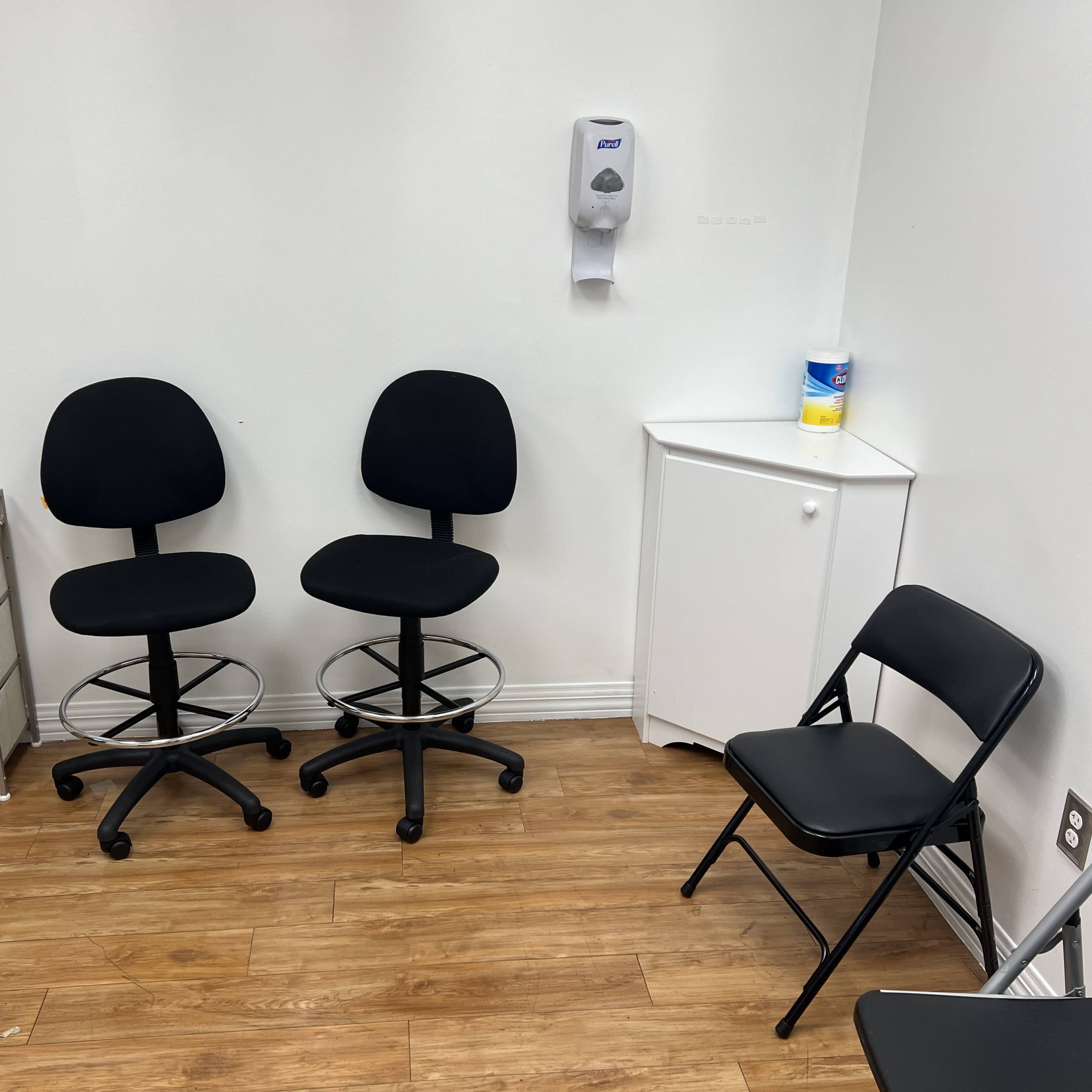 A small room with two black swivel chairs, a folding chair, a white cabinet, and a hand sanitizer dispenser on the wall.