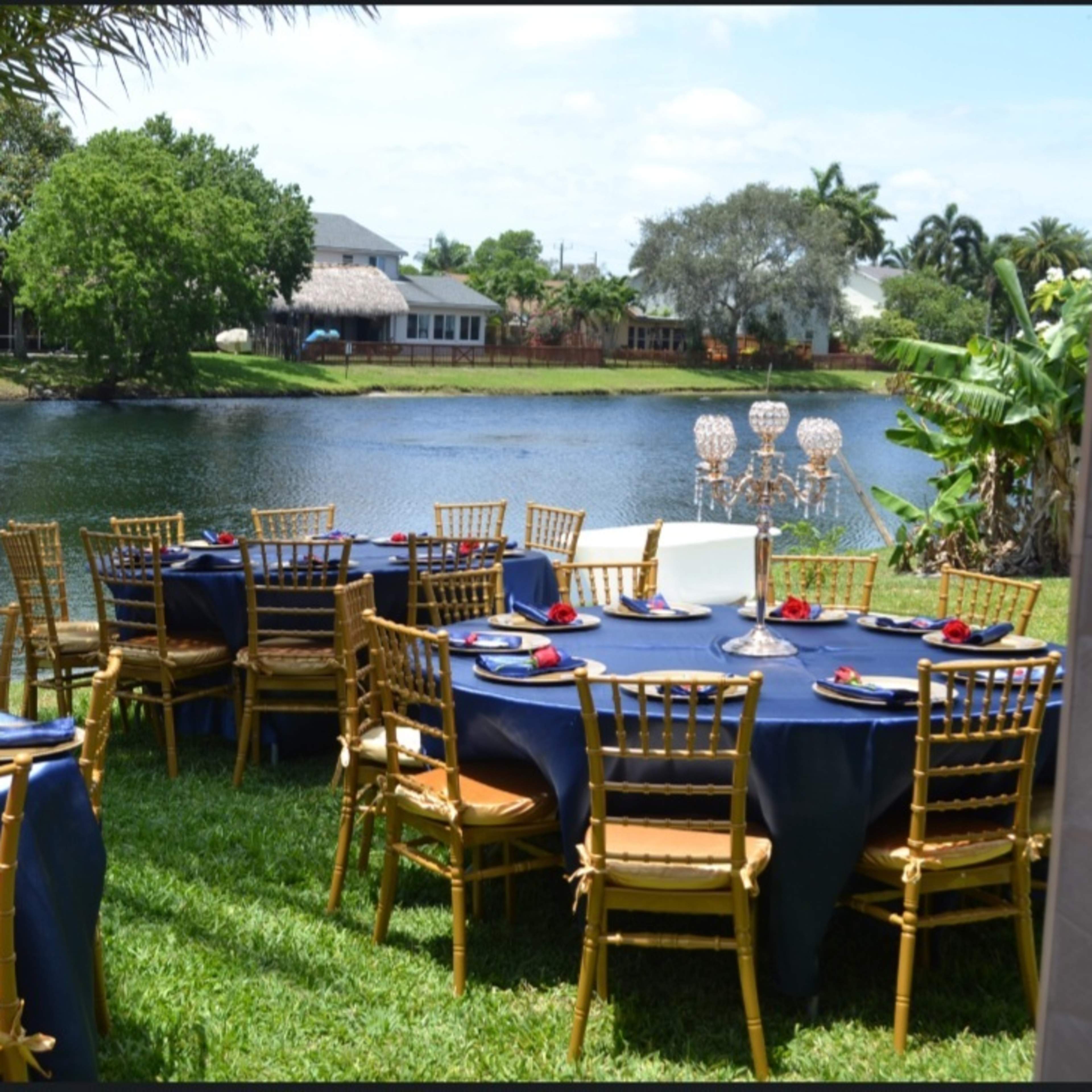 A beautifully arranged outdoor dining setup with golden chairs and blue tablecloths overlooks a serene pond.