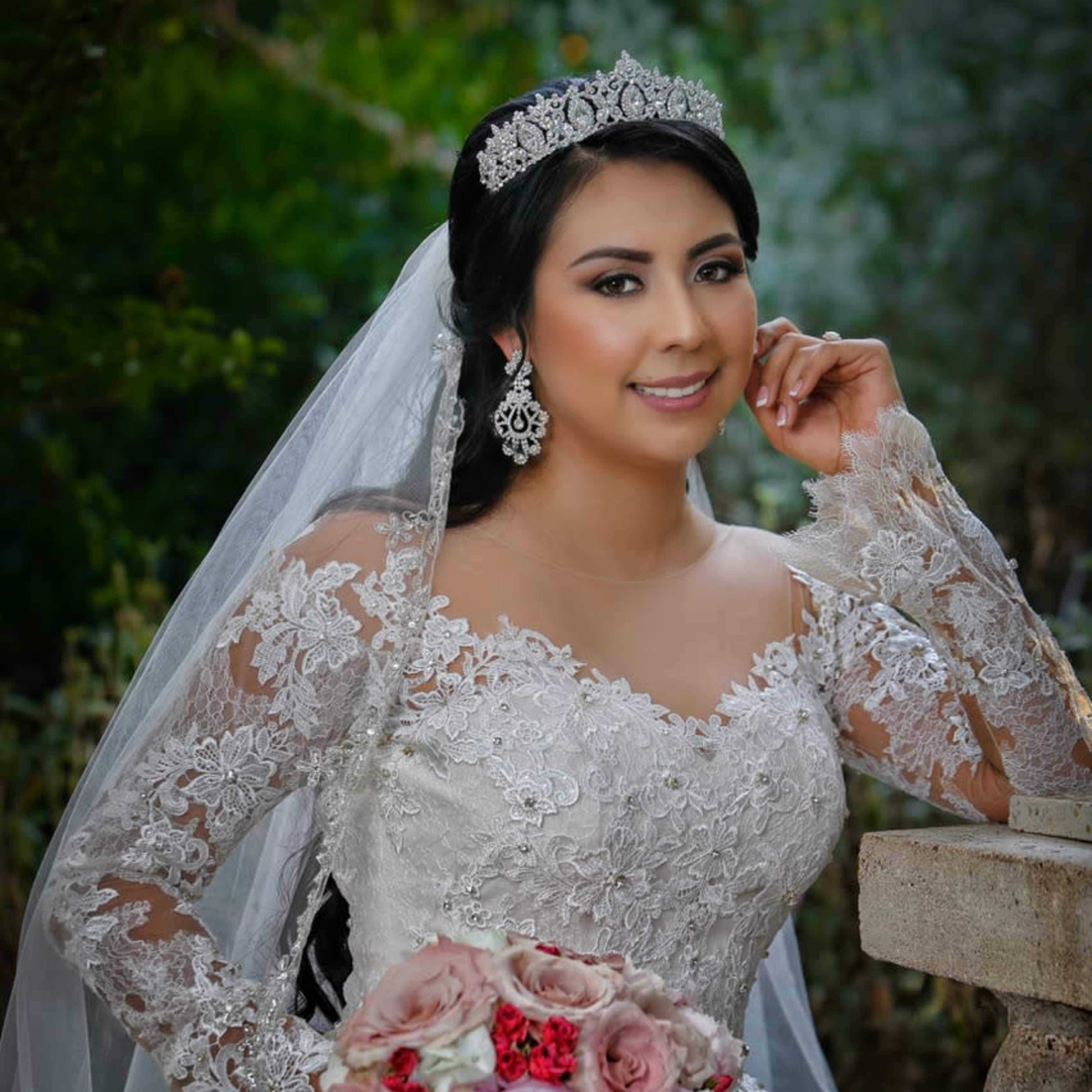 A bride in an ornate wedding dress poses with a bouquet of flowers near a stone pillar.