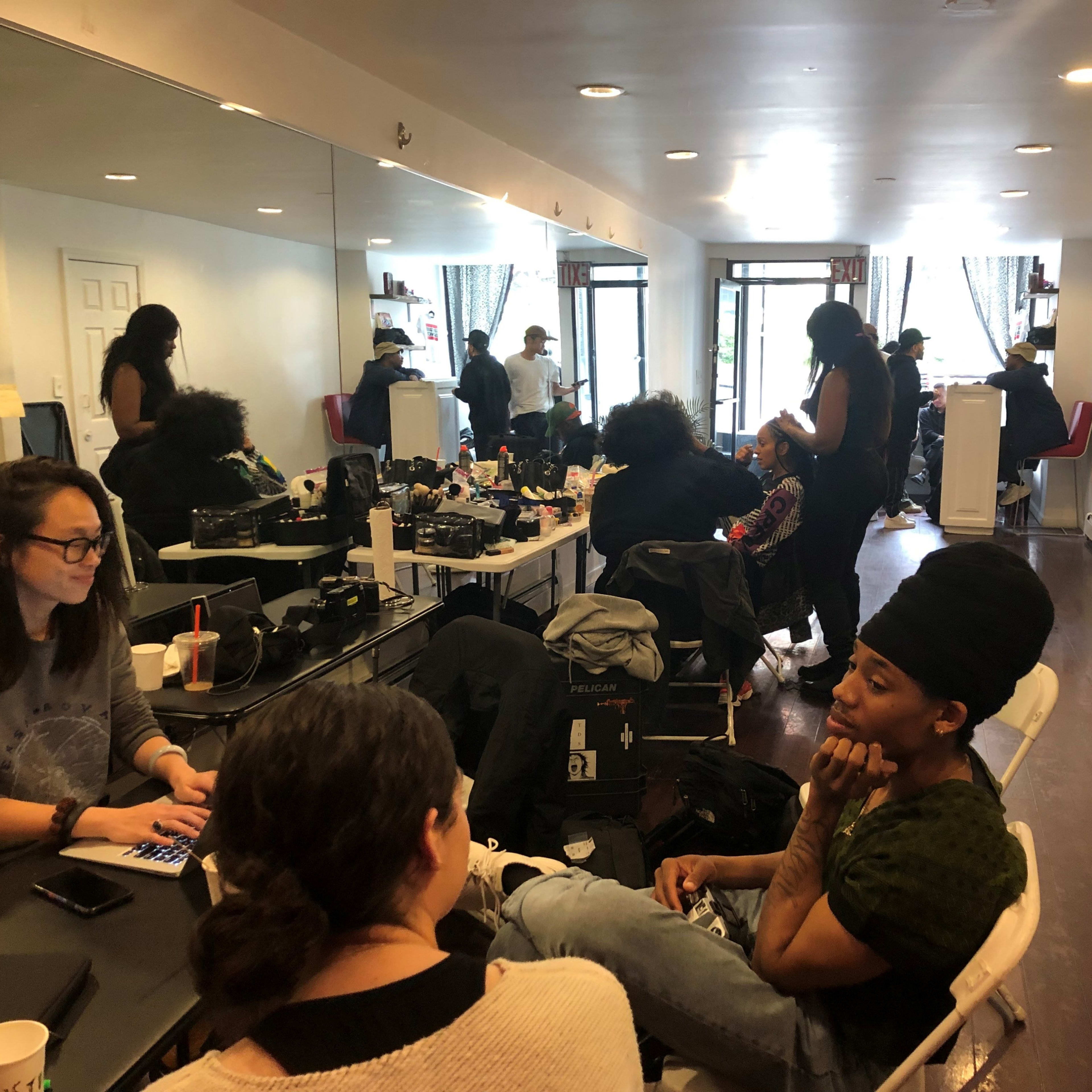 A group of people are engaged in hairstyling and conversation in a brightly lit salon with mirrors along one wall.