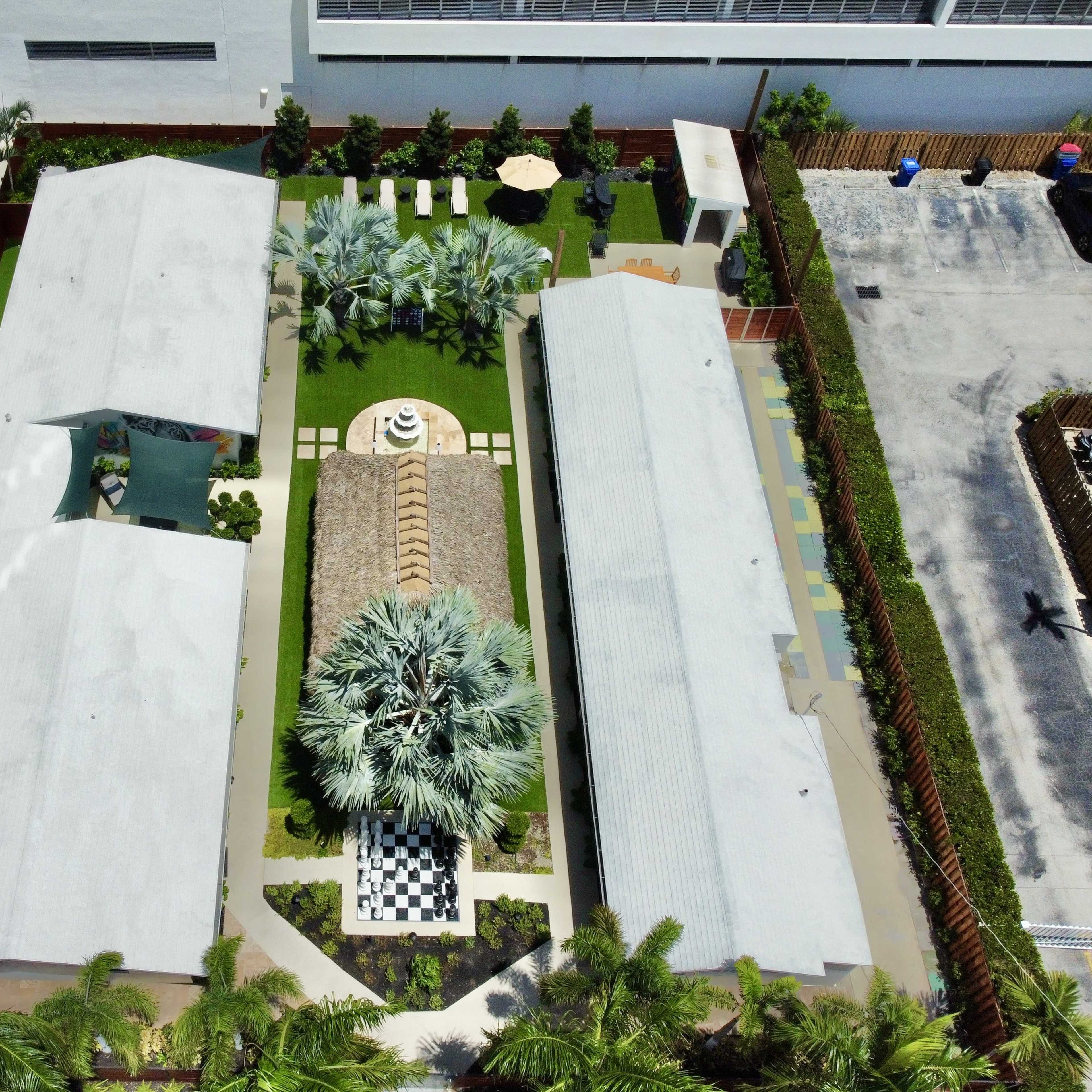 An aerial view of a landscaped area featuring two buildings flanking a central garden with a checkerboard path and a thatched gazebo.