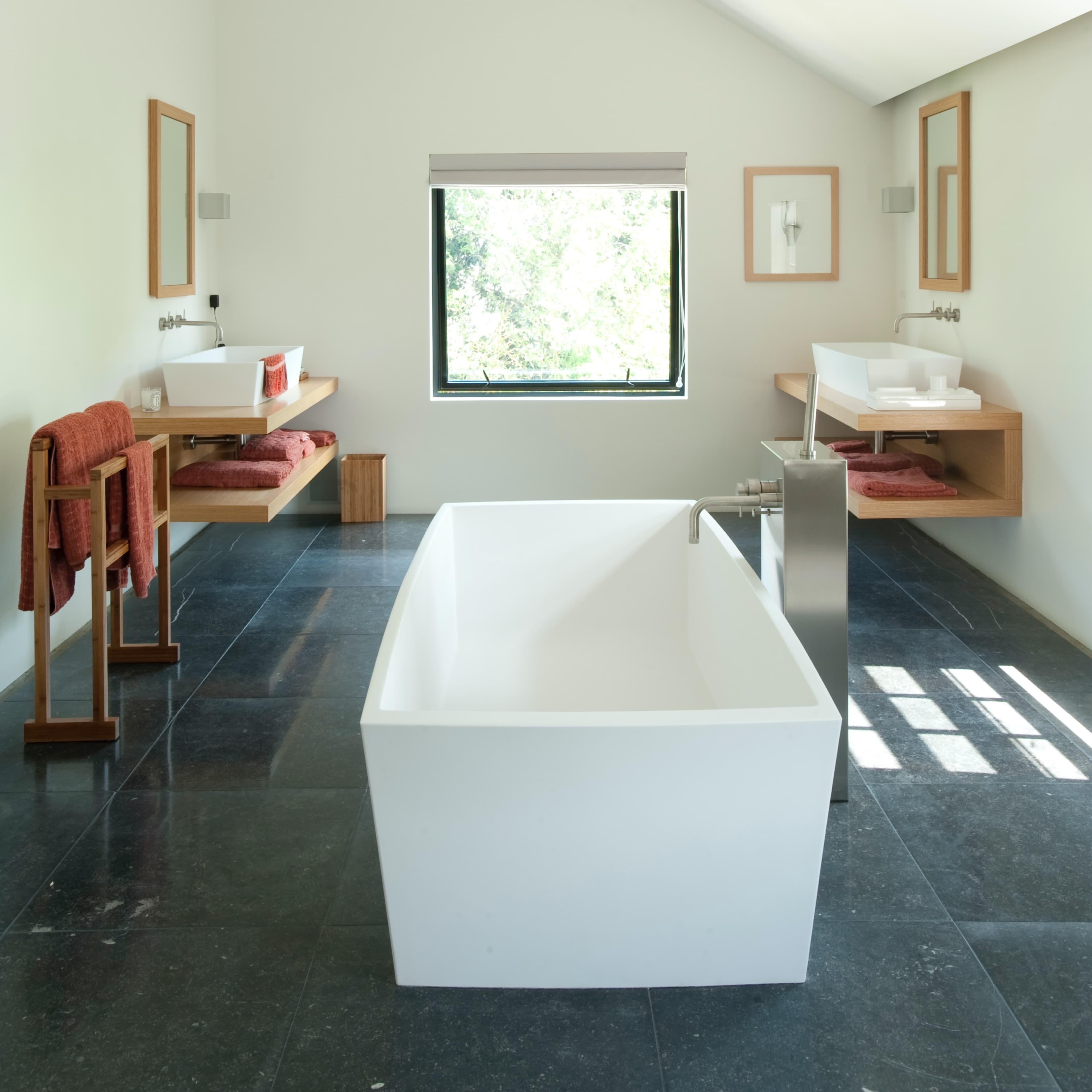 A modern bathroom featuring a freestanding white bathtub, a window with a view, and minimalist furnishings on stone flooring.