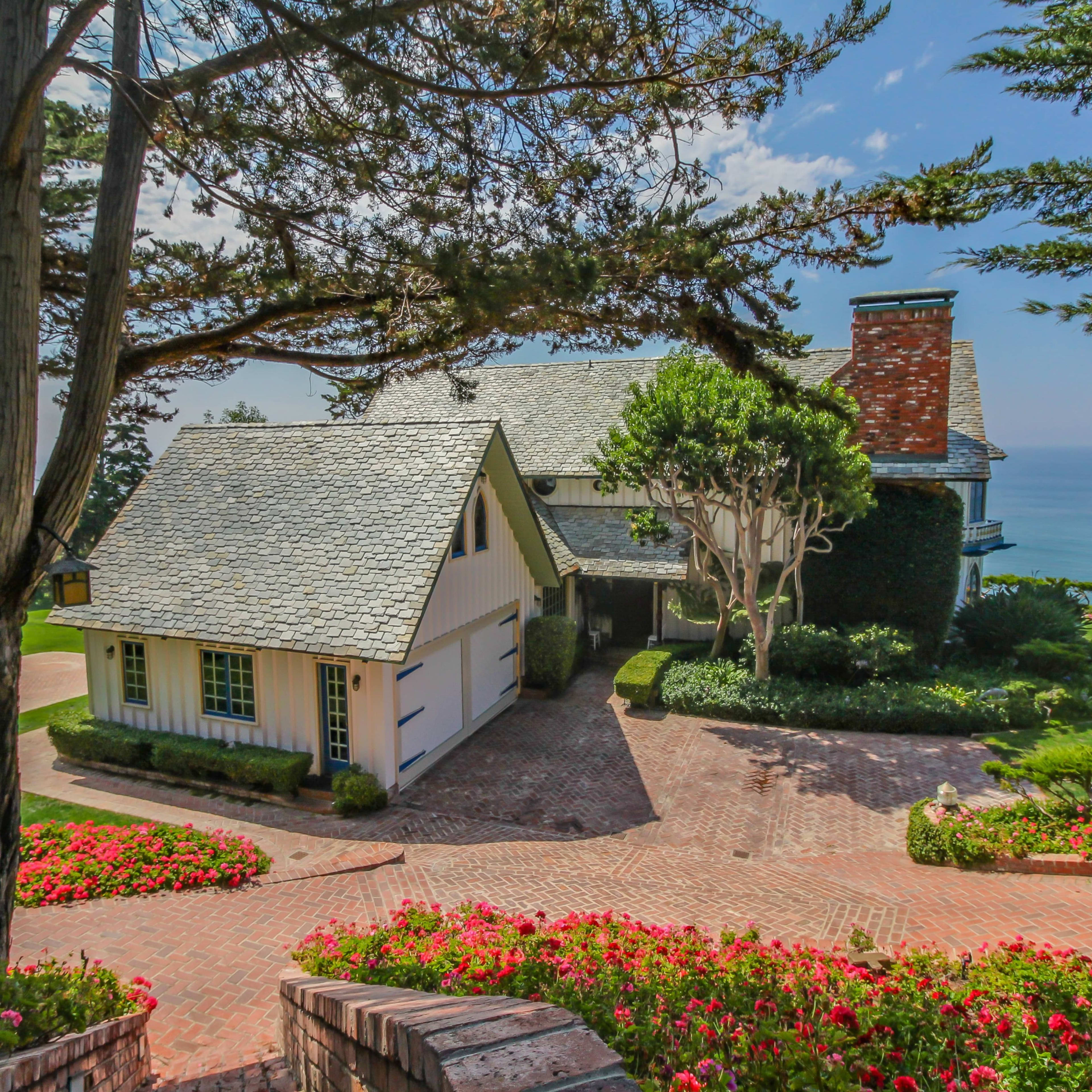 A large coastal house with a shingled roof is surrounded by lush greenery and flowers, overlooking the ocean.