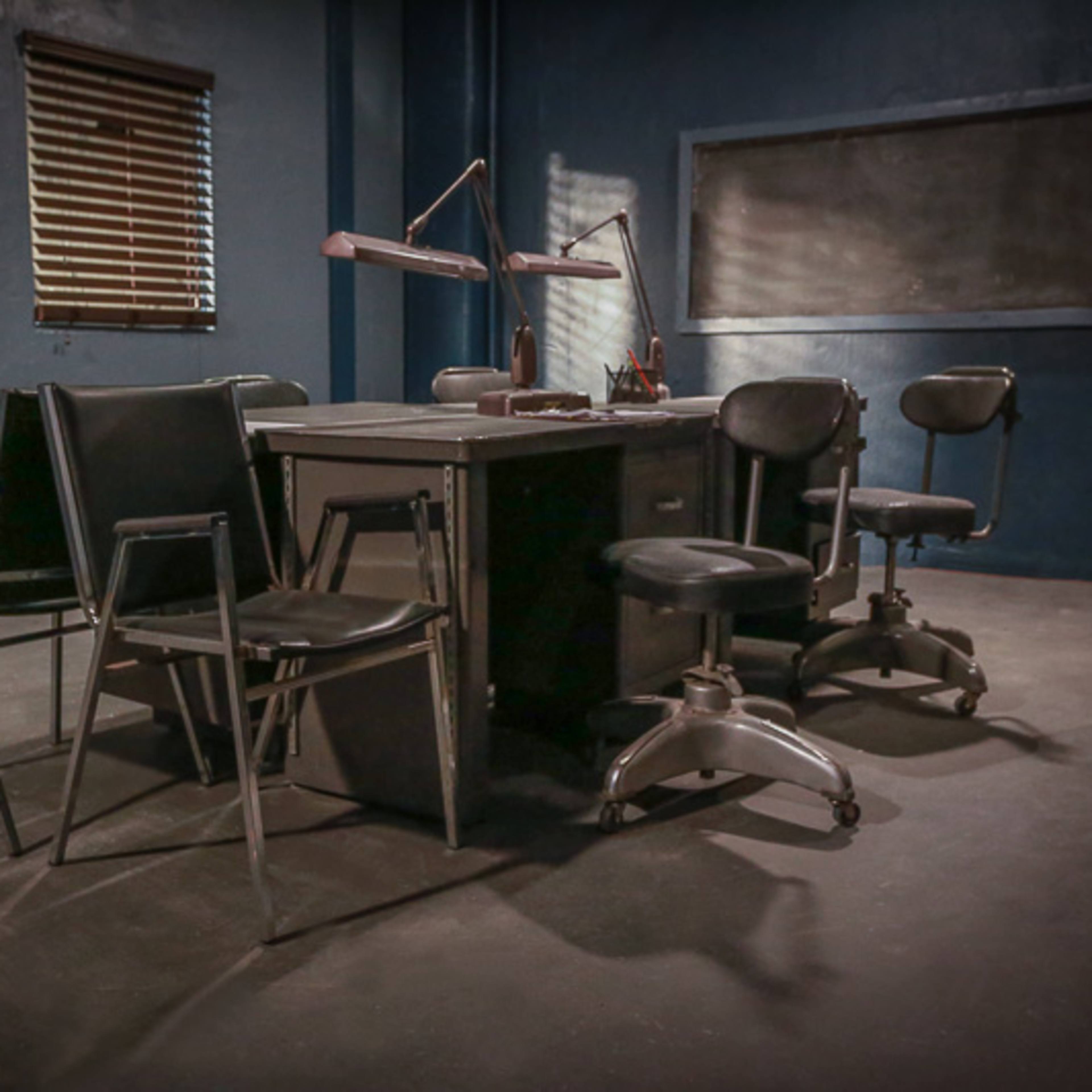 A dimly lit room features a table surrounded by several metal chairs and a couple of task chairs, with wooden blinds casting shadows on the walls.