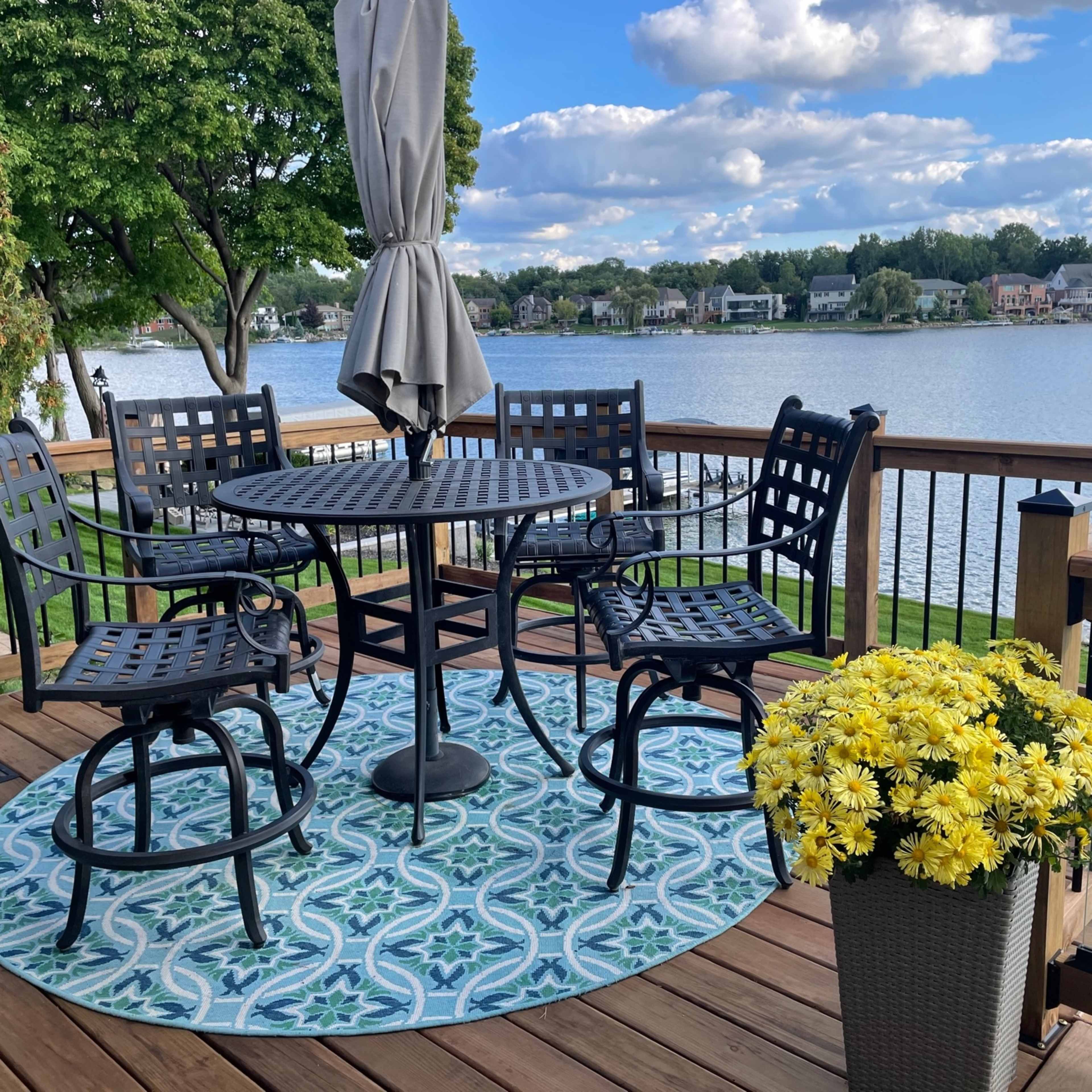 A round table with four chairs is set on a wooden deck overlooking a lake, surrounded by greenery and a potted plant filled with yellow flowers.