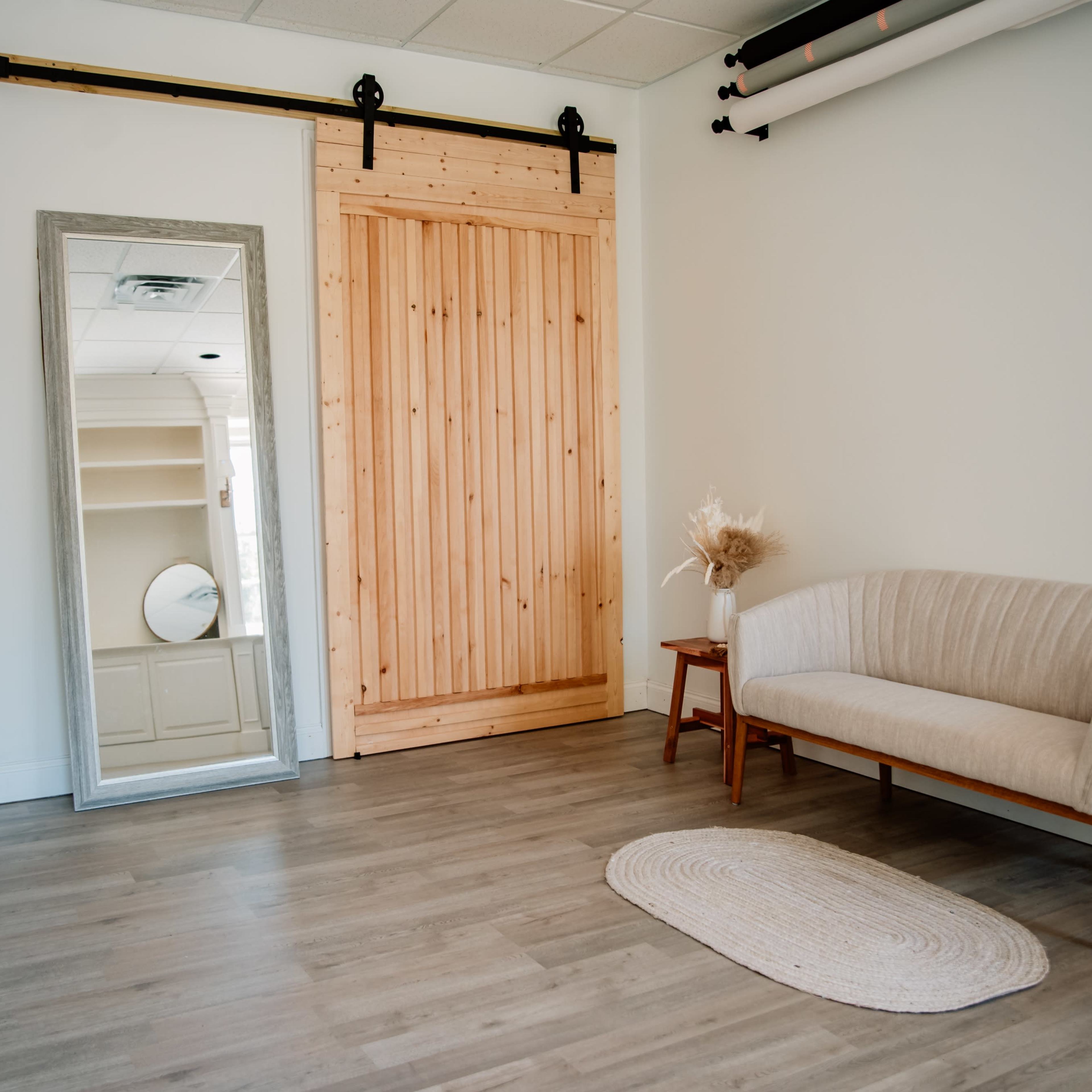 A minimalist interior space featuring a wooden sliding door, a full-length mirror, a light-colored couch, a small side table, and a round rug on a wooden floor.