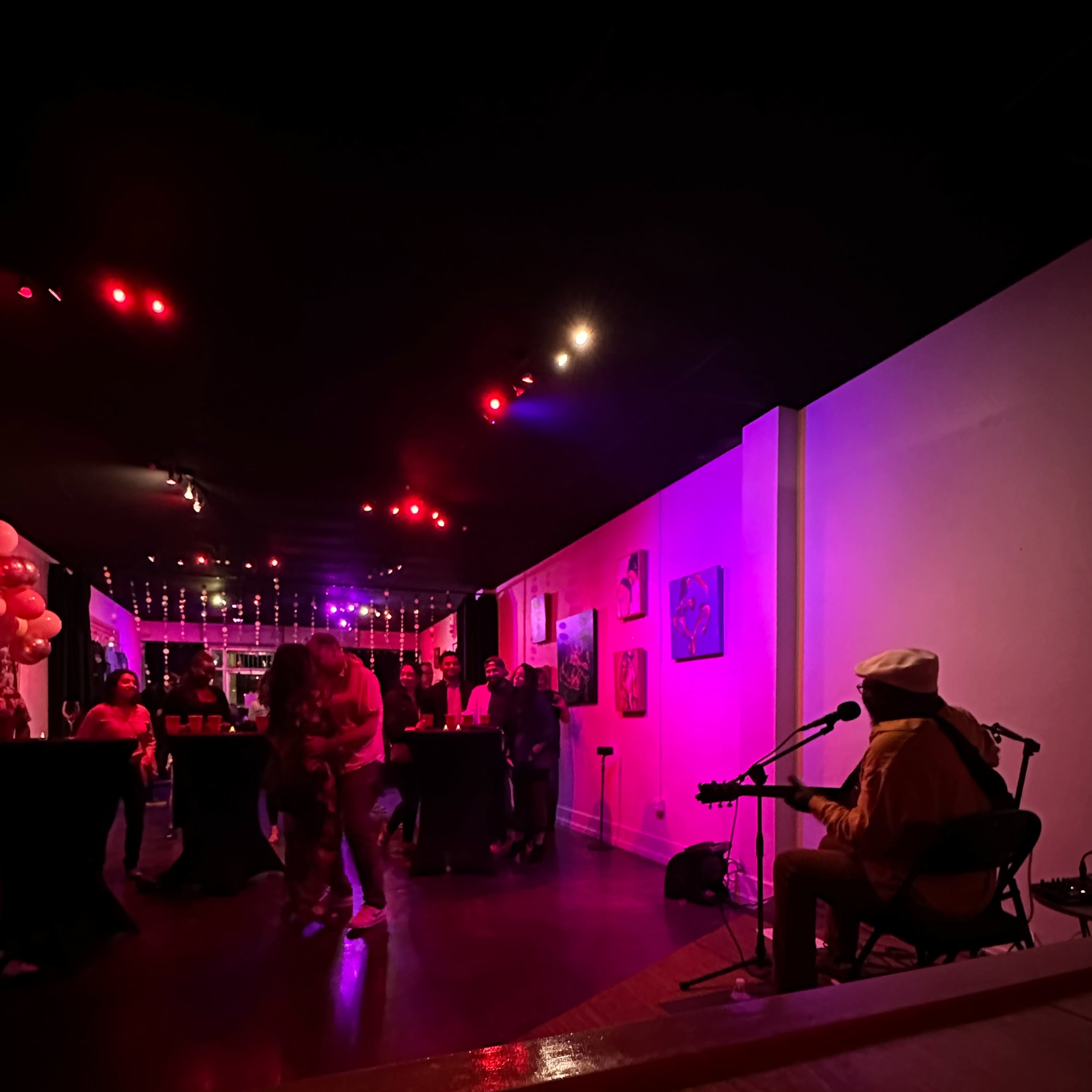A musician plays guitar in a dimly lit venue with guests dancing and socializing around tables decorated with balloons.