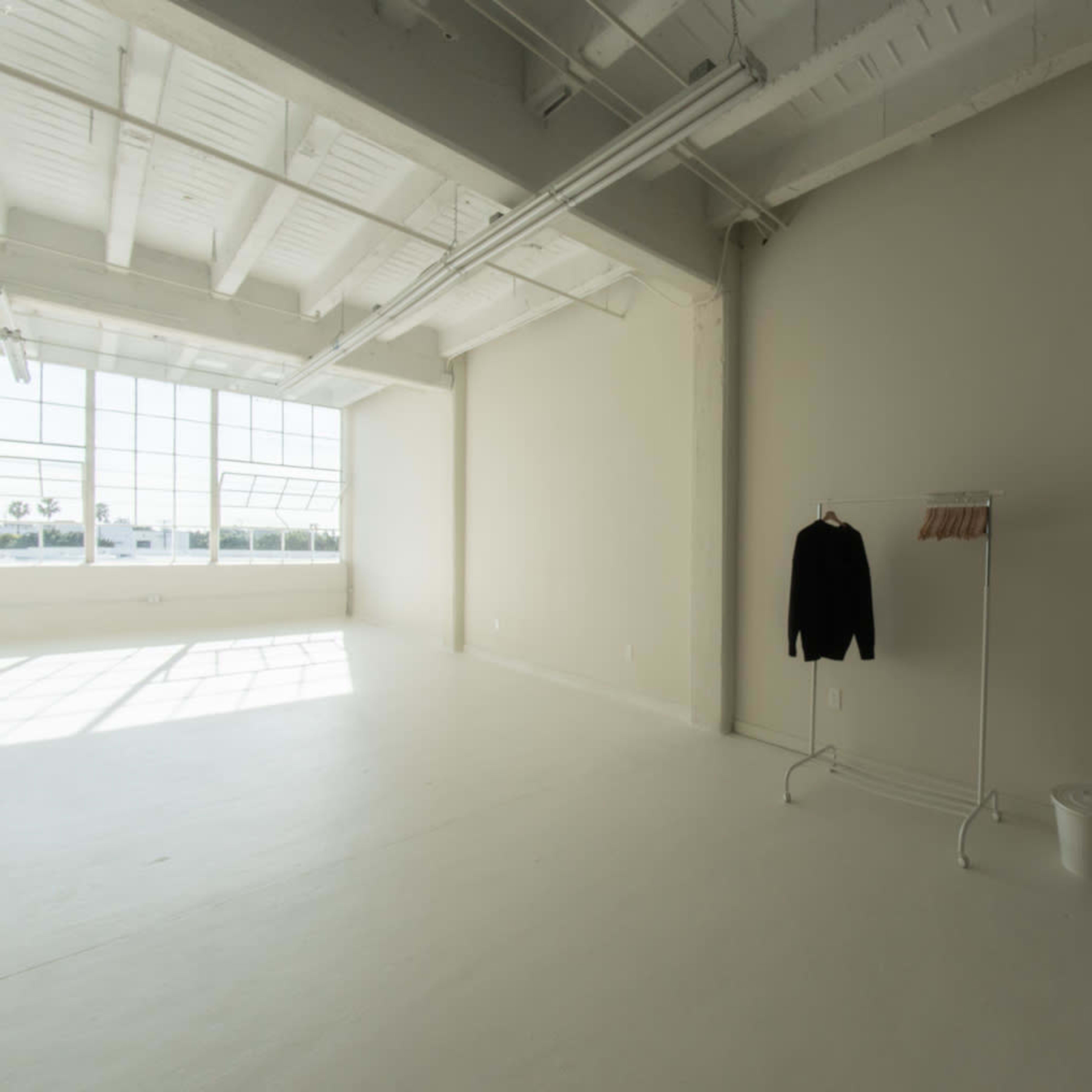 A spacious, empty room with white walls and a large window, featuring a clothing rack with a black garment, a white trash bin, and a black shelving unit.