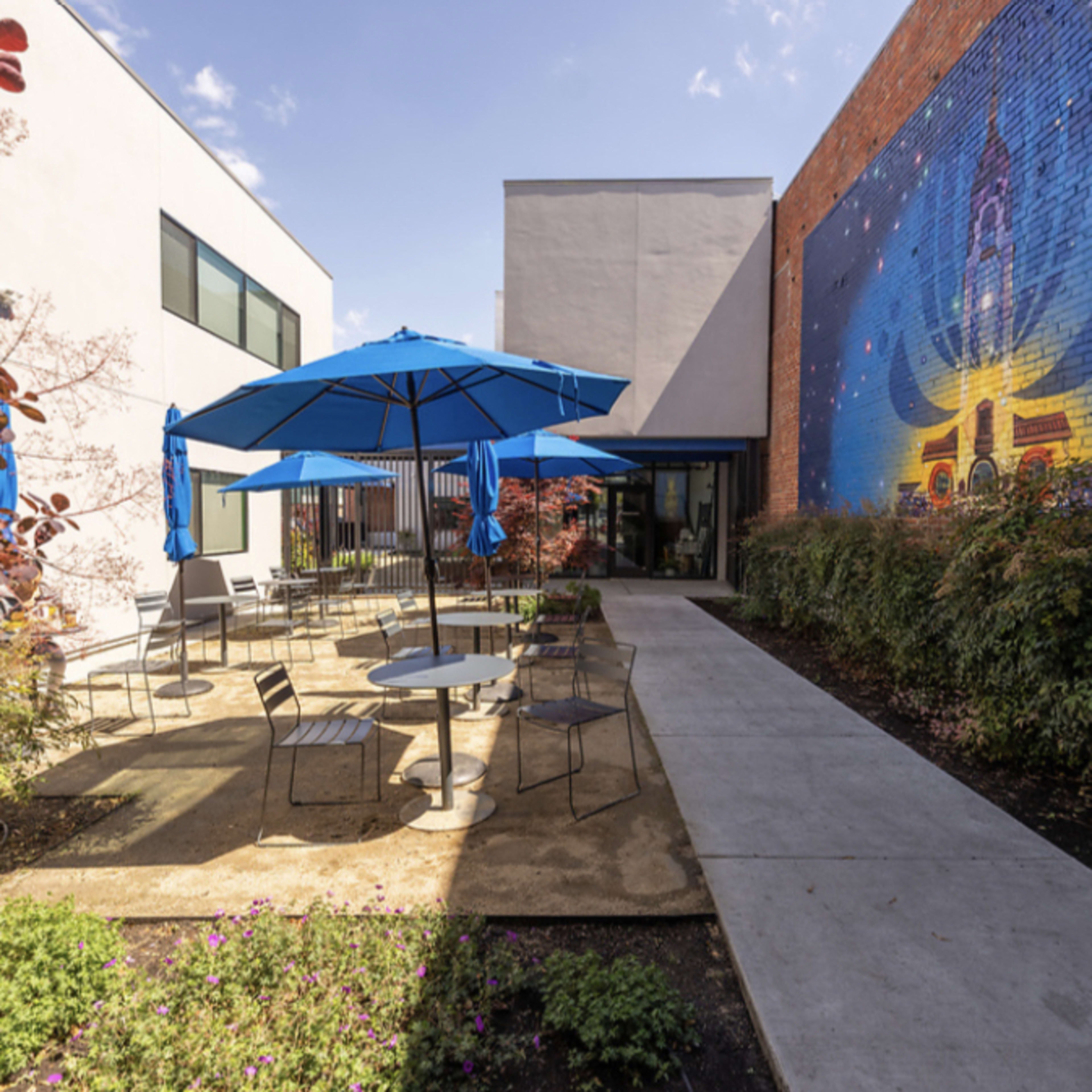 A courtyard with several blue umbrellas over round tables surrounded by greenery and a mural on the wall.
